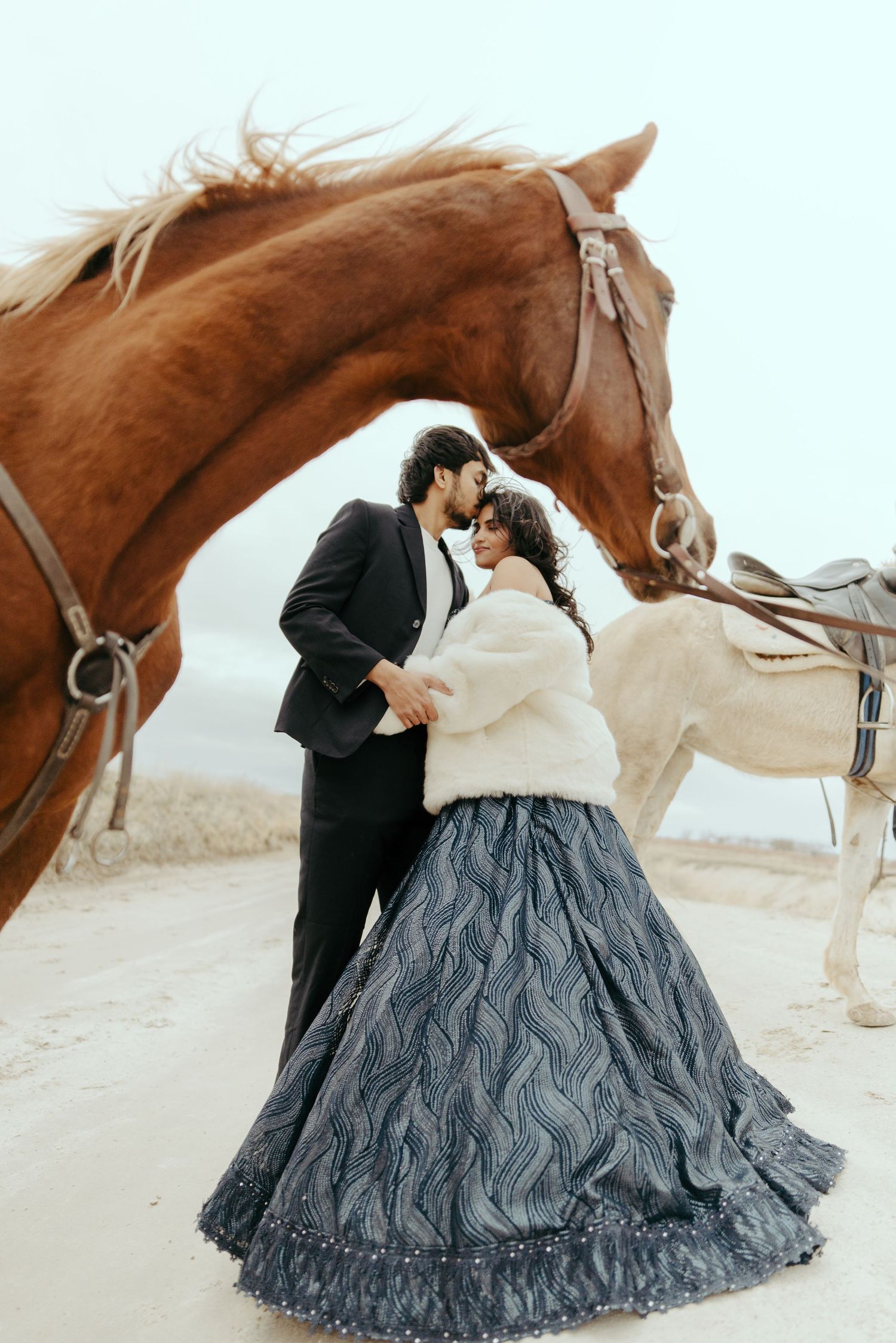 Couple embraces between brown and white horses, woman in blue lehenga and fur wrap.