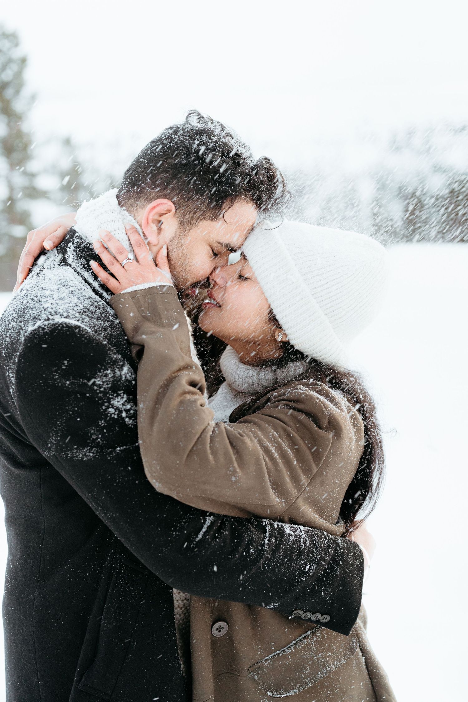 Romantic couple embracing and kissing in snowy winter landscape, wearing warm coats and hats covered in snow.