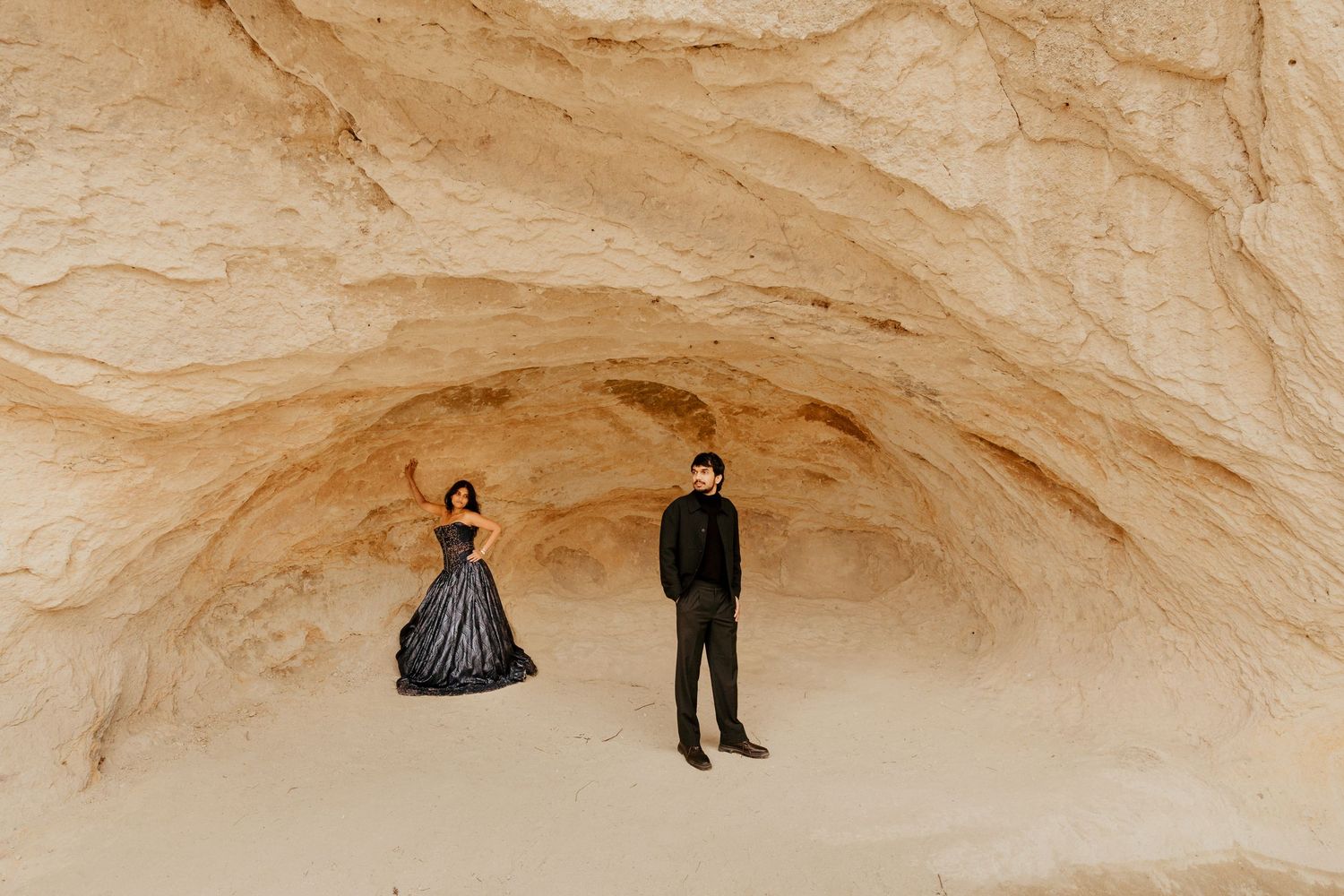 Couple in black wedding attire posing inside a sandstone cave formation for a dramatic desert elopement photo.