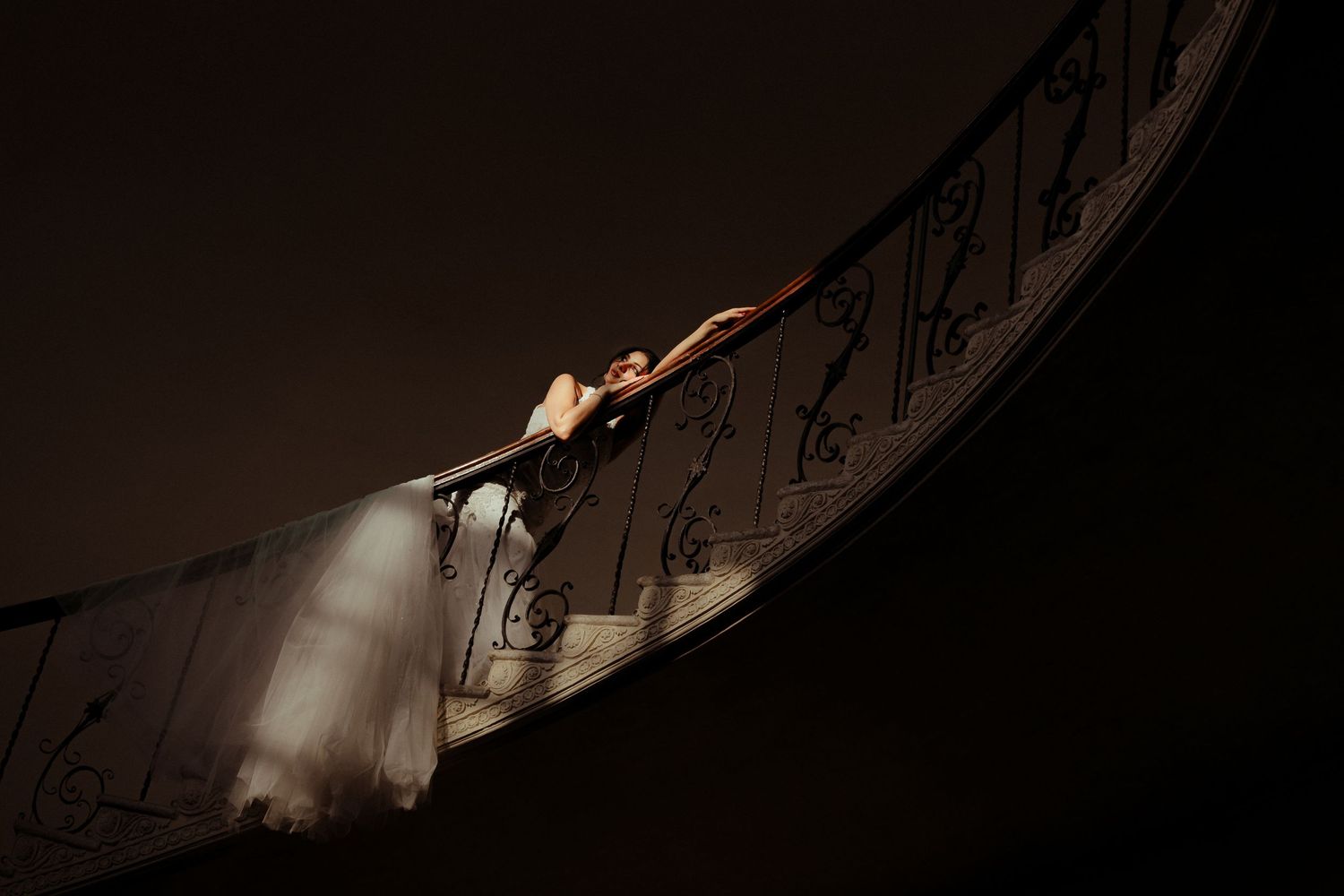 Bride in white gown poses gracefully on elegant curved staircase with ornate iron railing in dramatic dark lighting.
