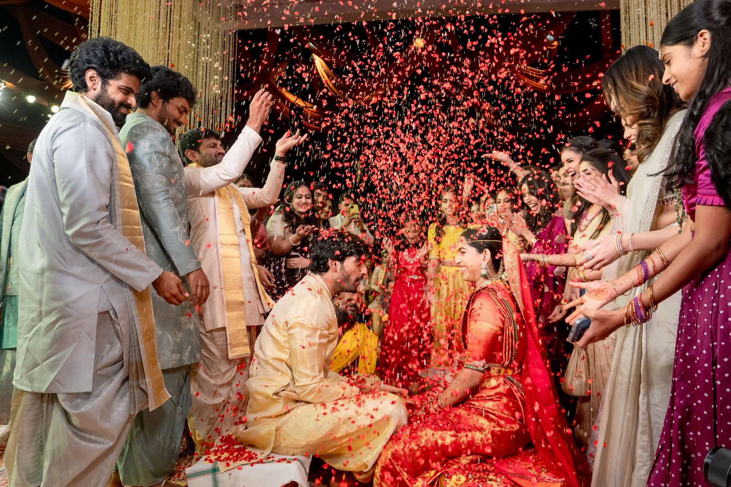 Traditional Indian wedding ceremony with bride in red saree and groom as guests shower colorful flower petals.