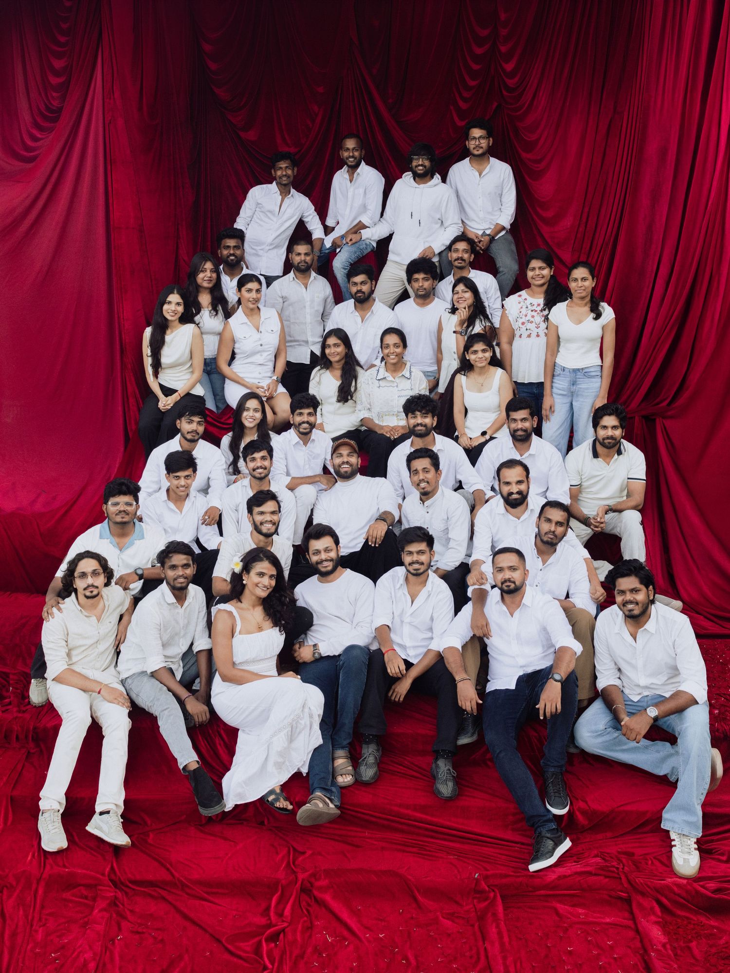Large group of people in white outfits posing together against a dramatic red curtain backdrop.