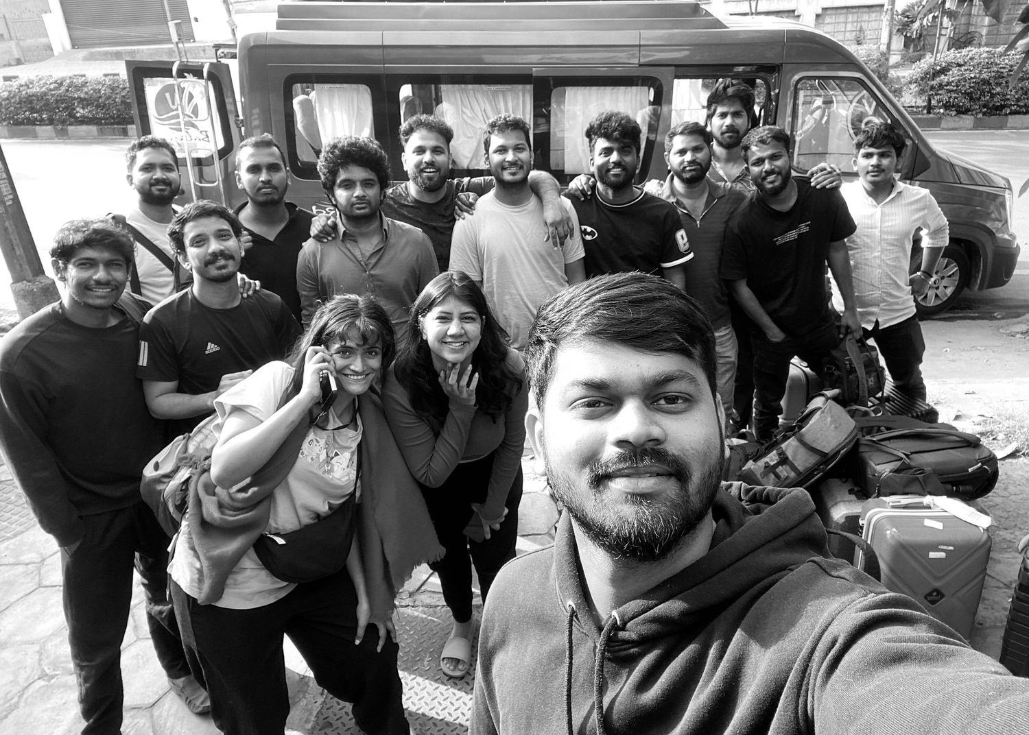 Black and white group selfie of diverse young adults posing outdoors near a van with bags and camera equipment.