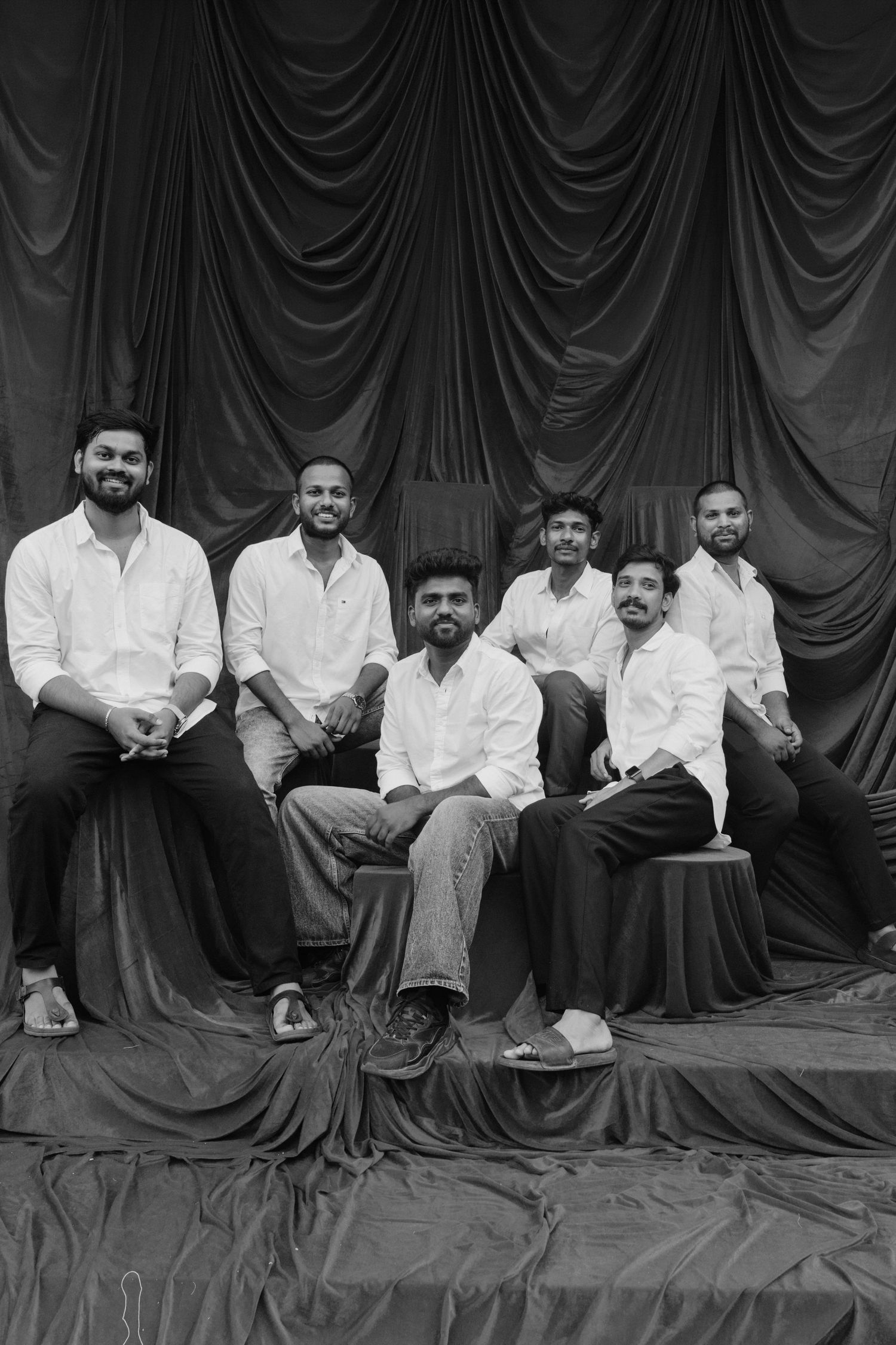 Six young men in white shirts pose together for a black and white group portrait against draped fabric backdrop.
