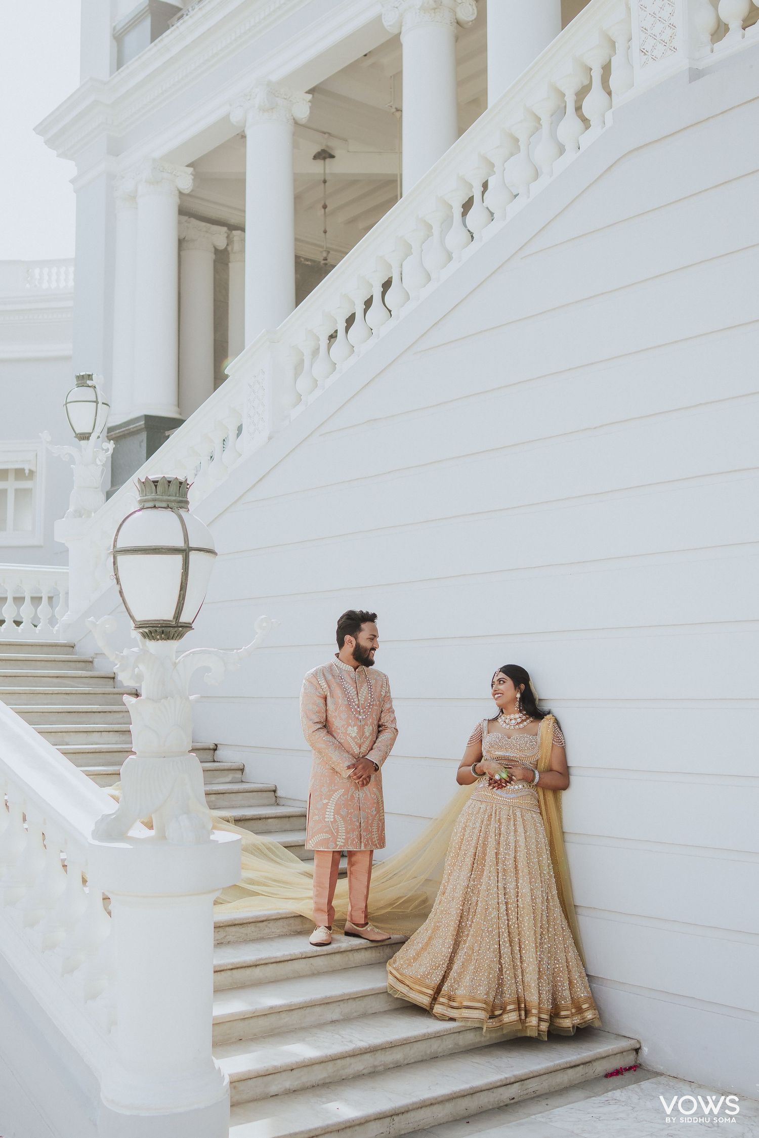 Indian couple in matching champagne gold outfits posing on white colonial building steps for wedding portrait.