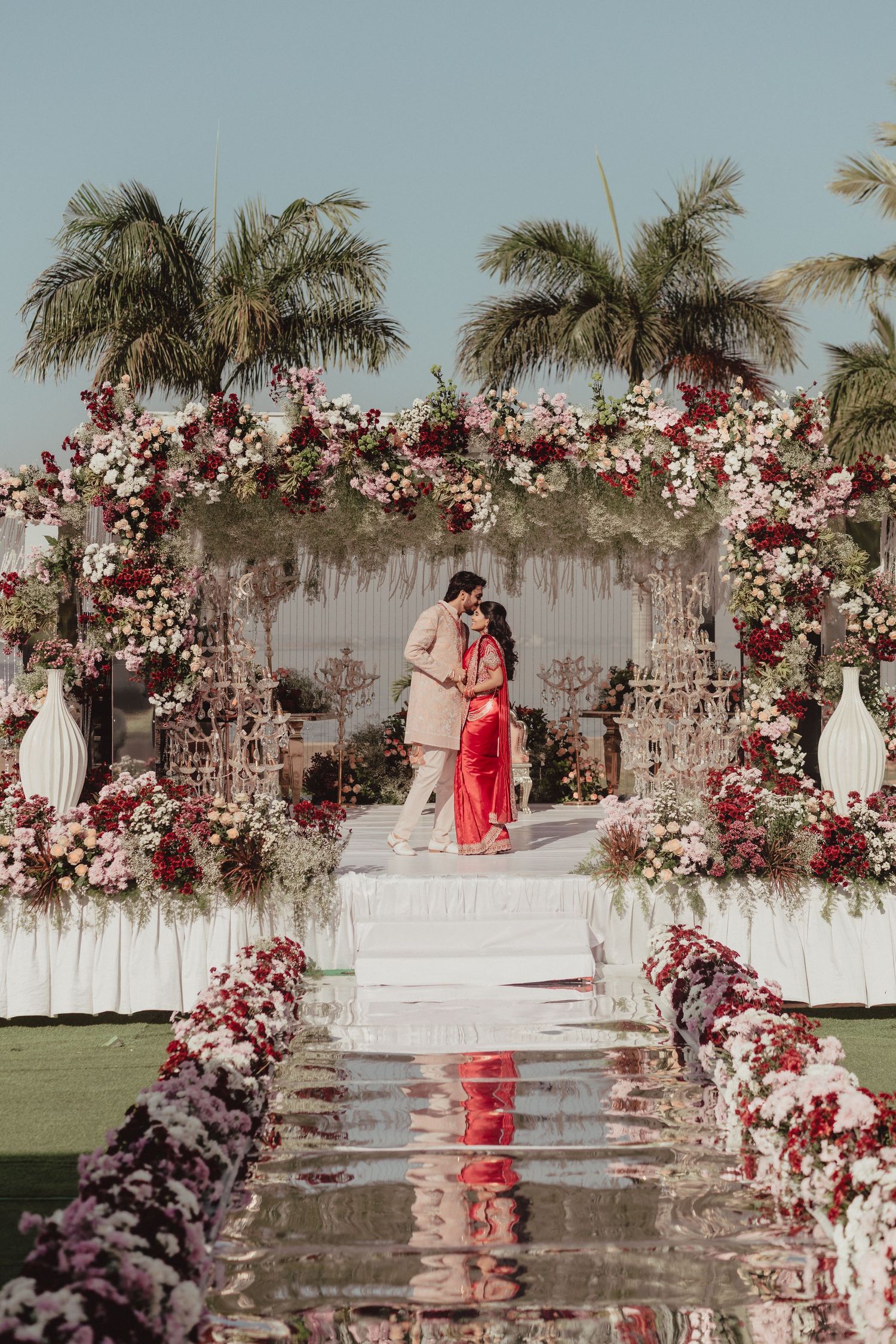 Indian wedding couple kissing under an elaborate floral arch with red and white blooms outdoors.