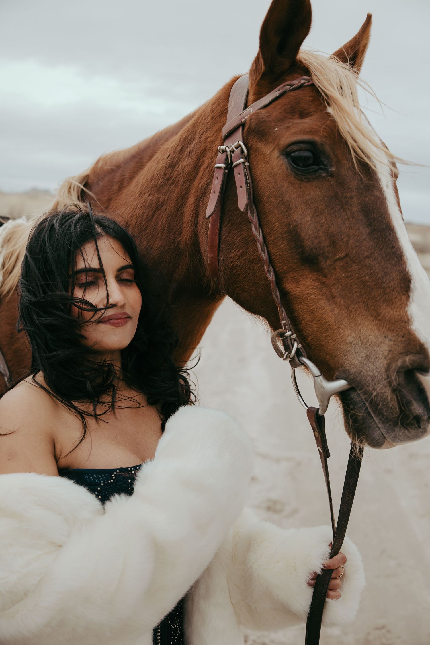 A woman in a white fur wrap holds the reins of a chestnut horse outdoors under a cloudy sky.