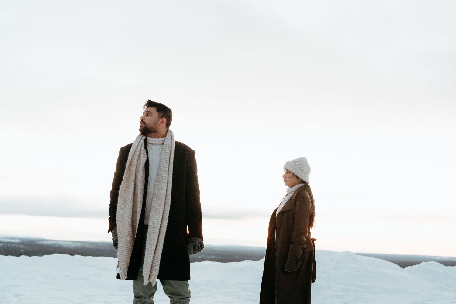 A man and woman in winter coats and scarves stand apart in a snowy landscape under a pale sky.