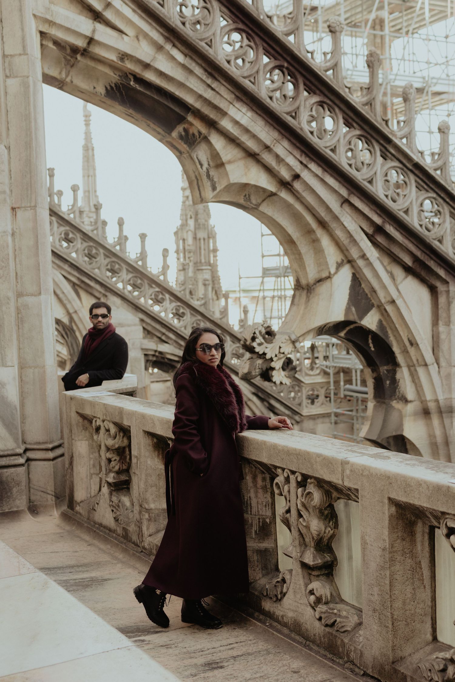 Woman in burgundy coat poses on ornate Gothic cathedral rooftop terrace with arches and spires.