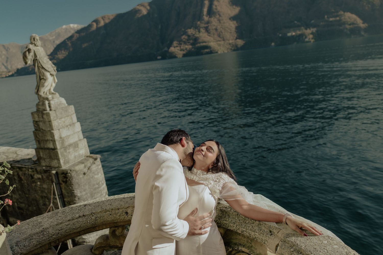 Couple sharing romantic kiss on ornate stone balcony overlooking Lake Como with mountain backdrop on wedding day.