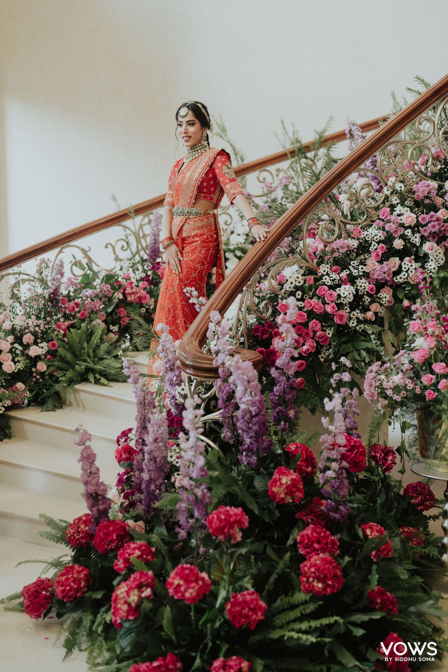 Indian bride in red lehenga poses on flower-adorned staircase with pink and purple blooms at wedding ceremony.