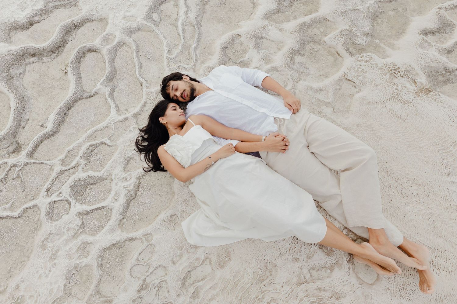 Couple in white outfits lying together on a white sandy beach, embracing in a romantic aerial wedding photo.