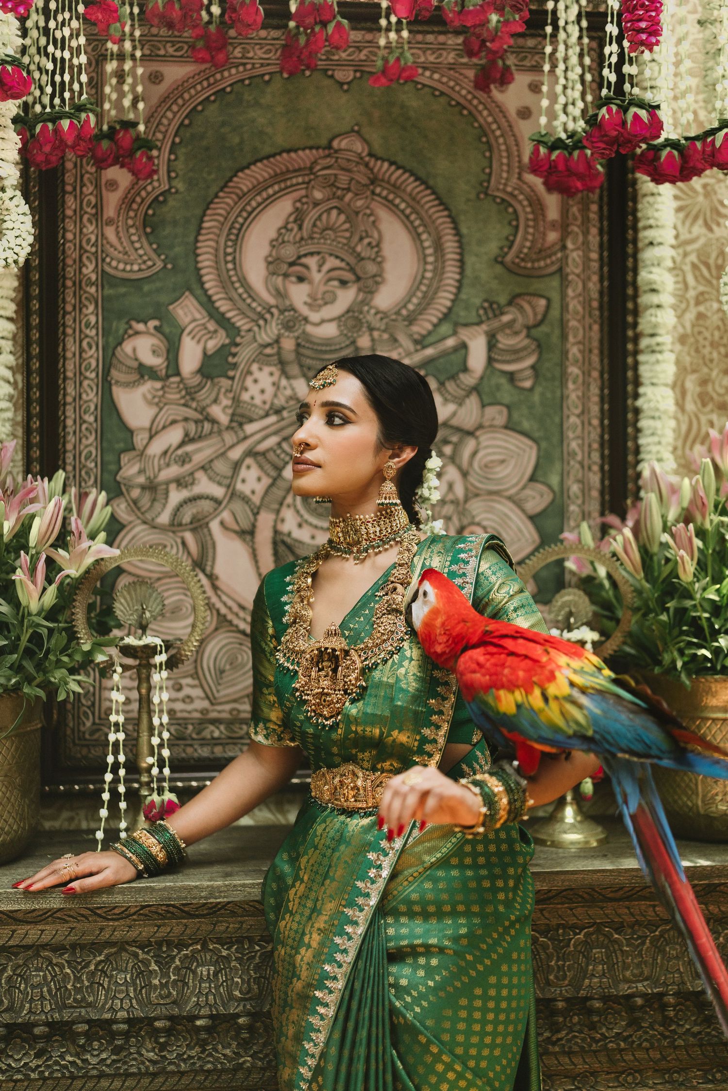 Woman in a green and gold silk saree with temple jewelry holds a red macaw beside lilies and a Saraswati painting.