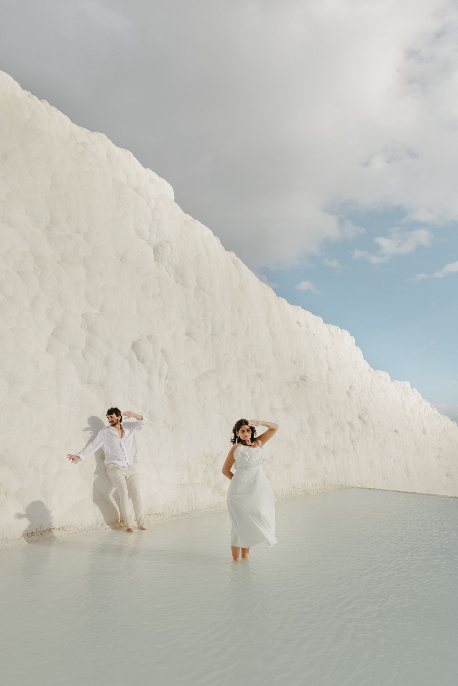 Couple in white outfits walks through Pamukkale travertine terraces with turquoise water.