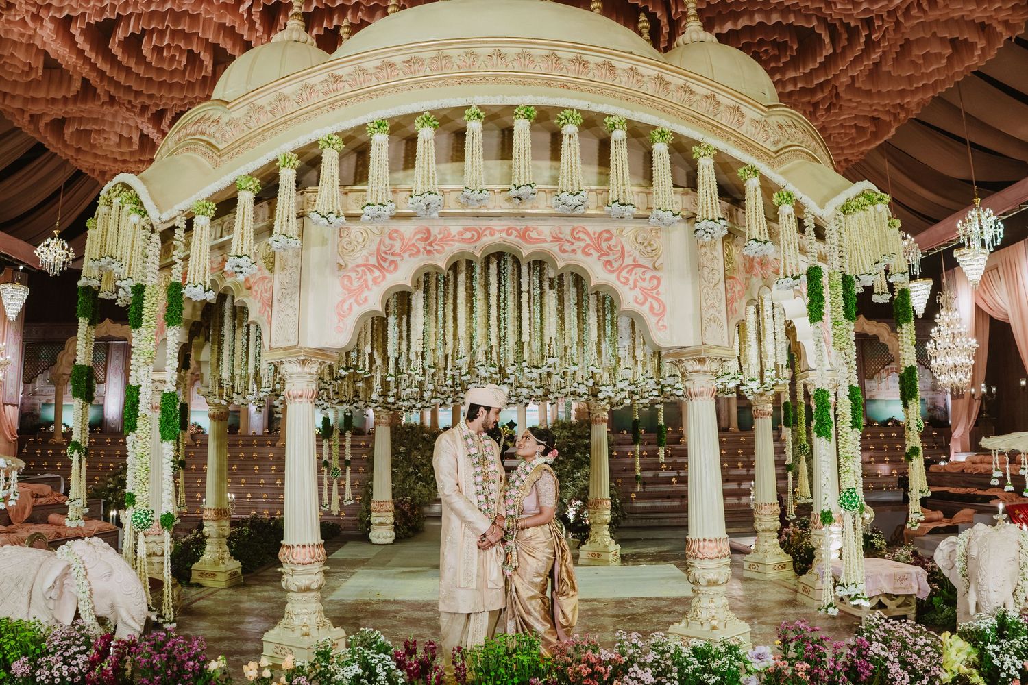 Couple stands beneath ornate mandap with floral decor at Indian wedding ceremony.