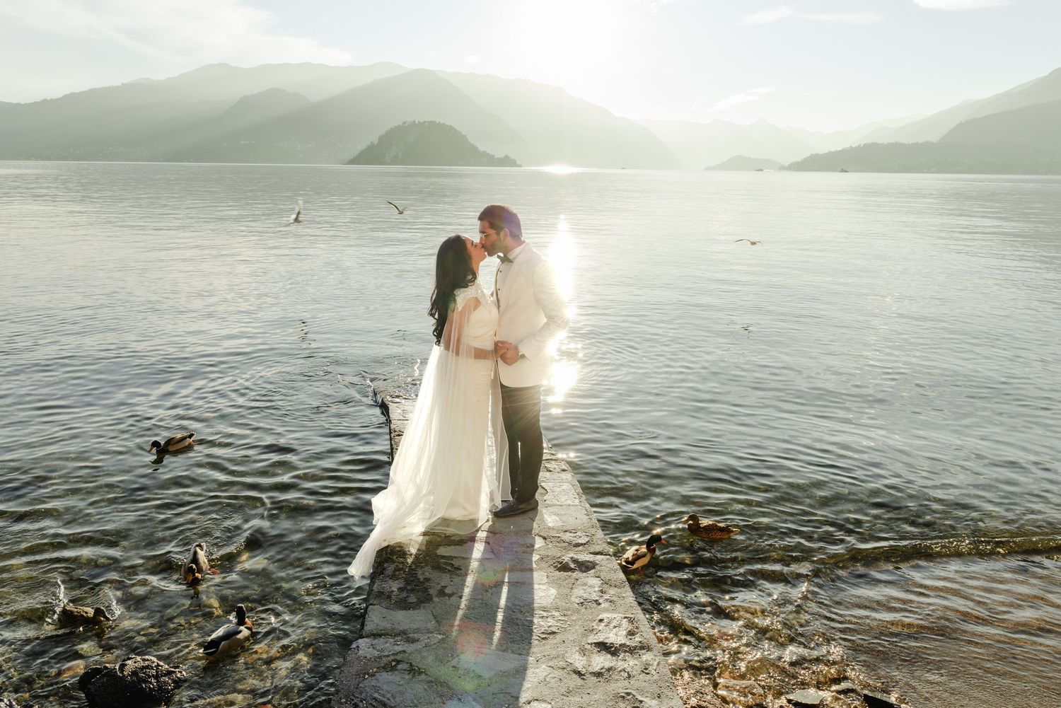Bride and groom kissing on lakeside pier surrounded by mountains and sparkling sunlight on calm water.
