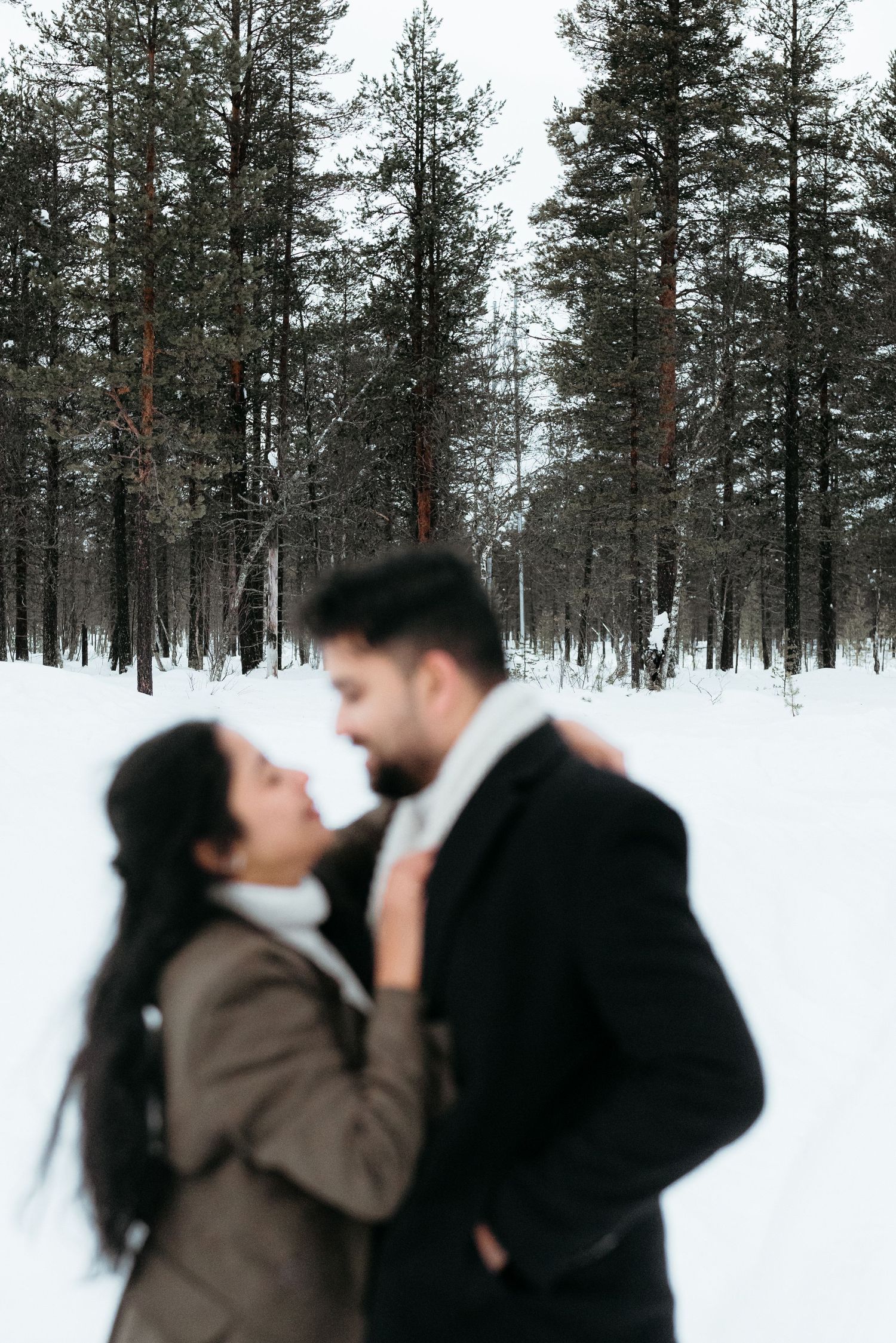 Couple embracing romantically in a snowy winter forest, surrounded by tall pine trees covered in snow.
