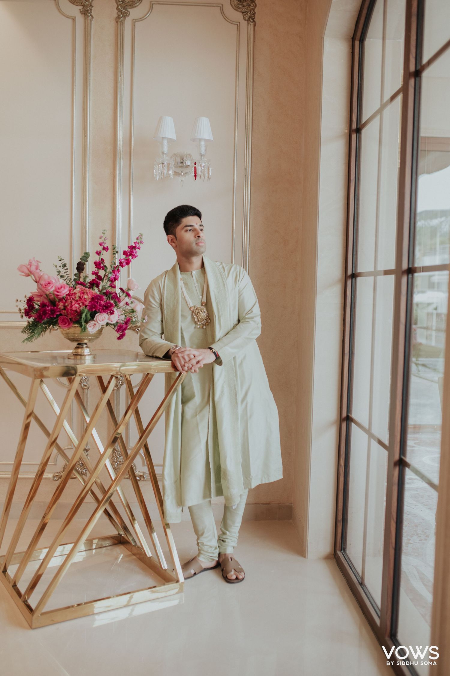 Groom in mint green sherwani poses by gold geometric table with pink floral arrangement in elegant venue.