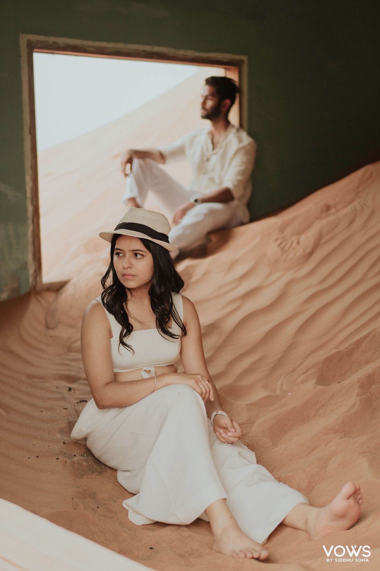 A woman in white outfit and hat sits on desert sand dunes while a man in white sits behind a green wall frame.