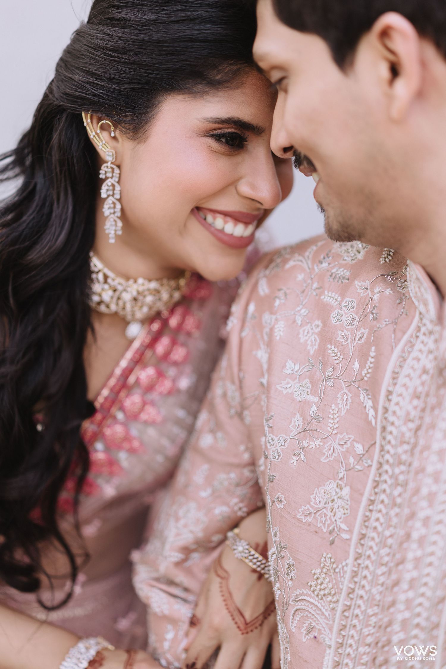 Indian couple in pink embroidered wedding attire sharing an intimate moment, bride wearing diamond jewelry.