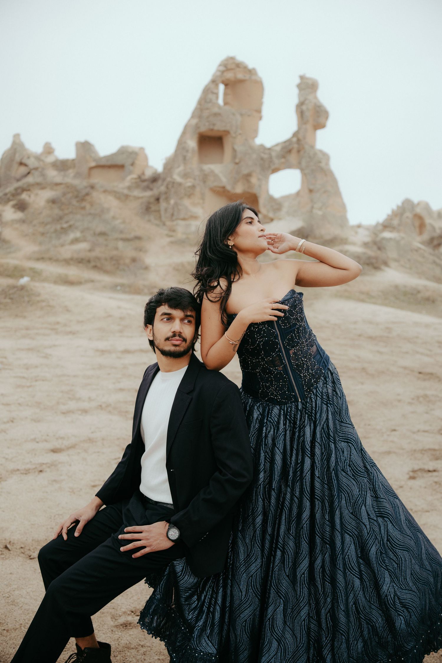 A couple poses dramatically in a desert landscape with ancient ruins, she wears a stunning navy blue gown.