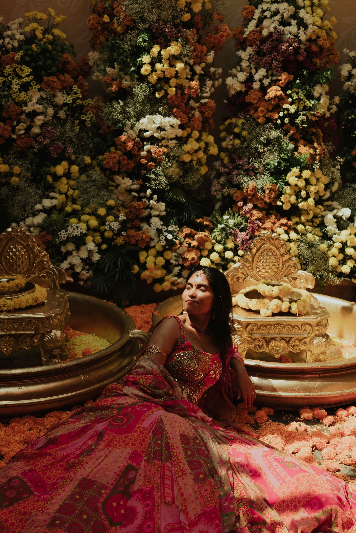 Bride in vibrant red lehenga seated among elaborate floral decorations at Indian wedding.