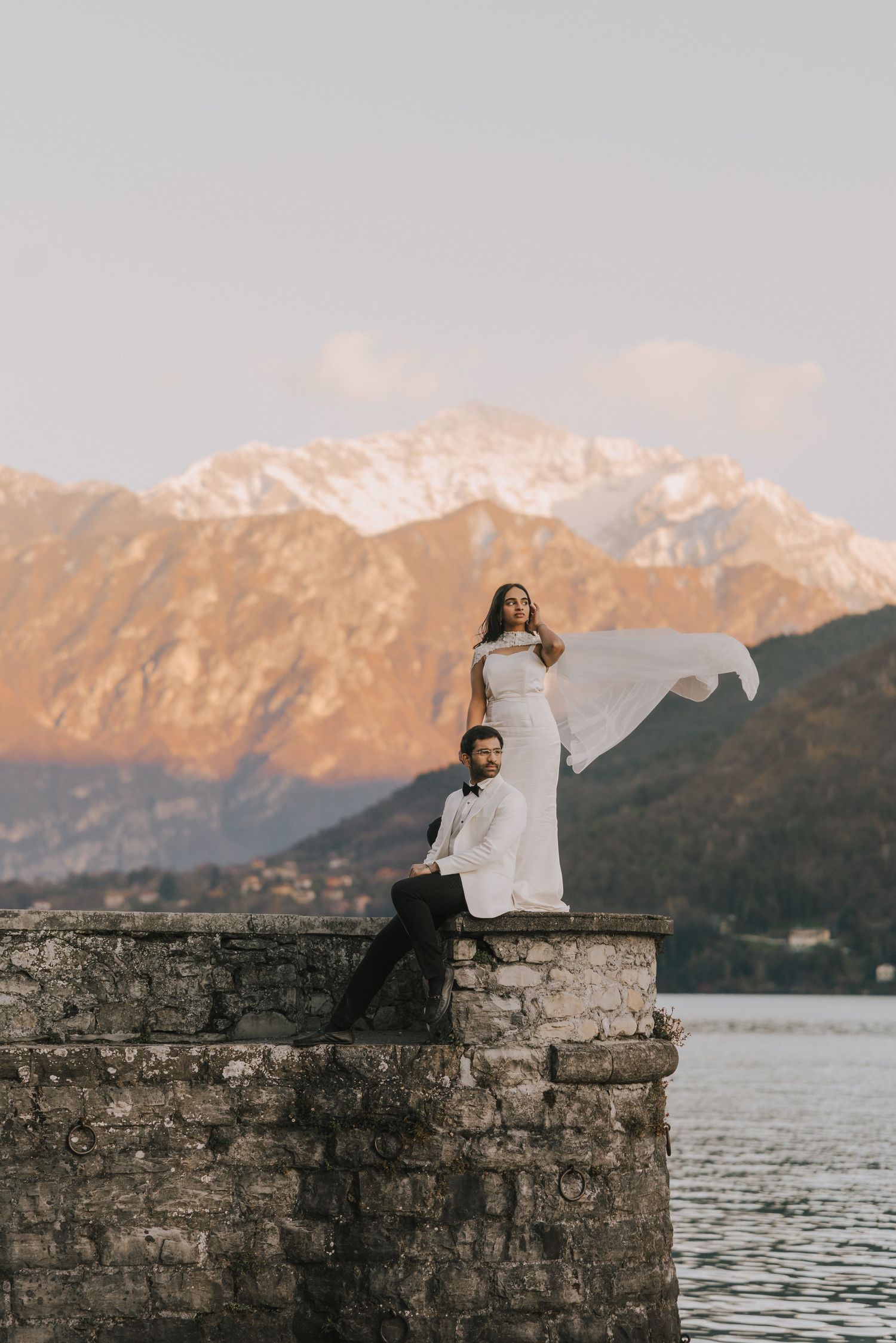 Wedding couple poses on stone wall by alpine lake, bride's dress flowing in wind at sunset.