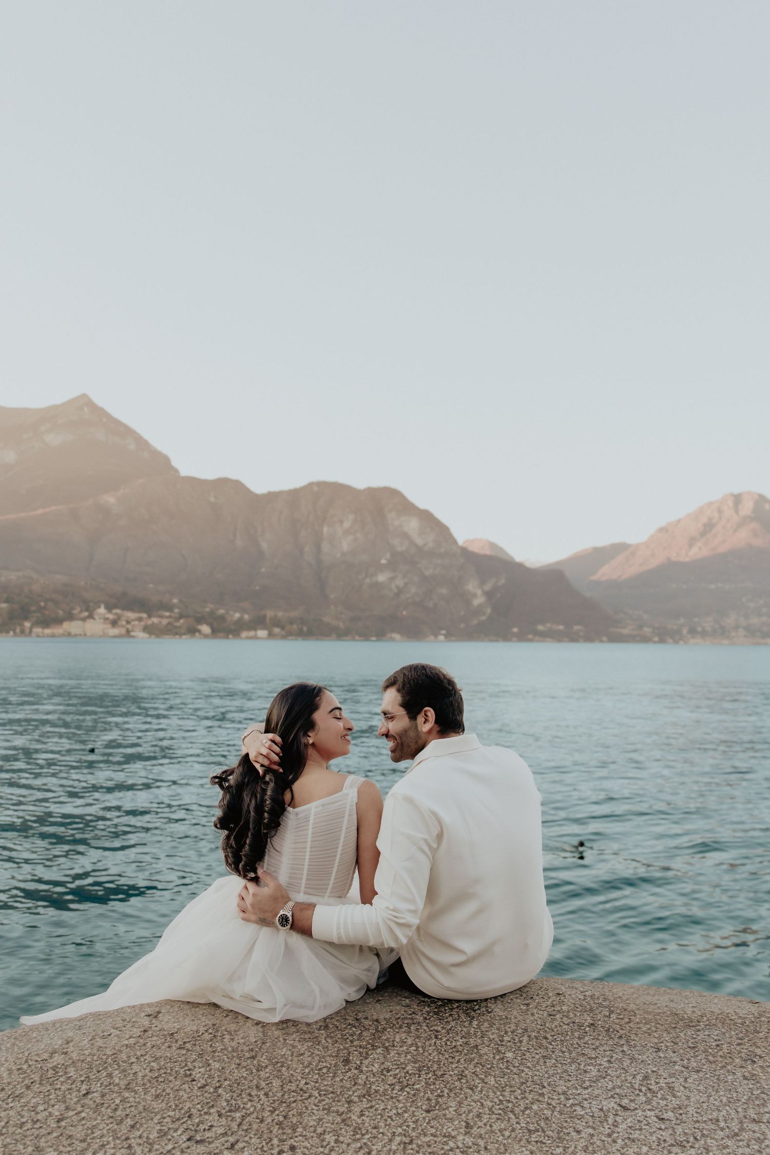 Couple in white attire sitting on a lakeside ledge with mountain backdrop at golden hour.