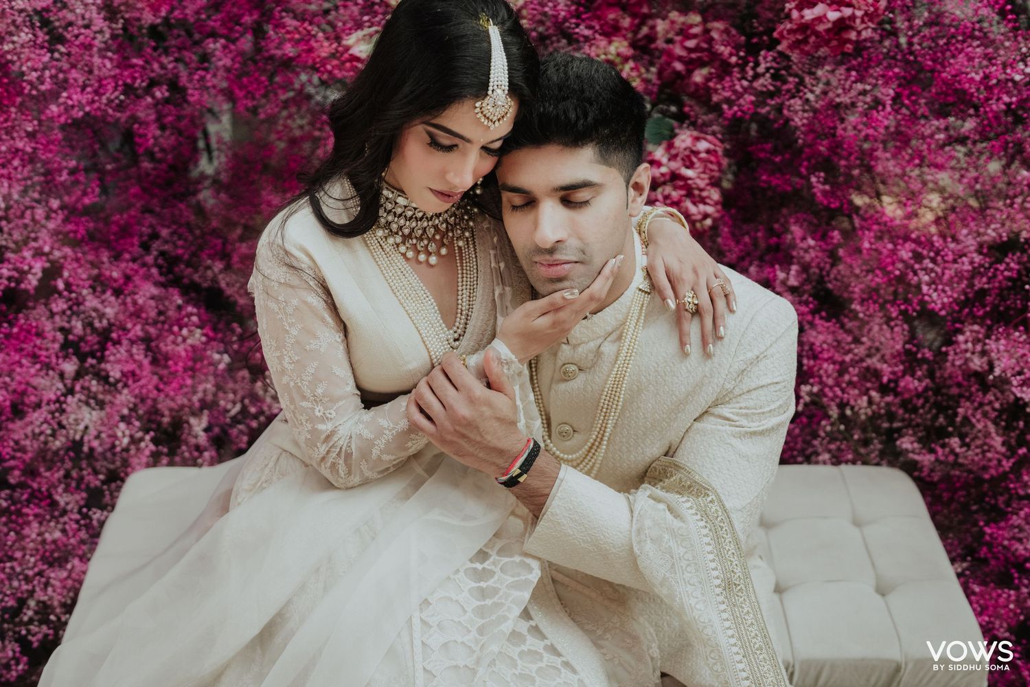 Couple in white Indian wedding attire posing romantically against vibrant pink bougainvillea floral backdrop.