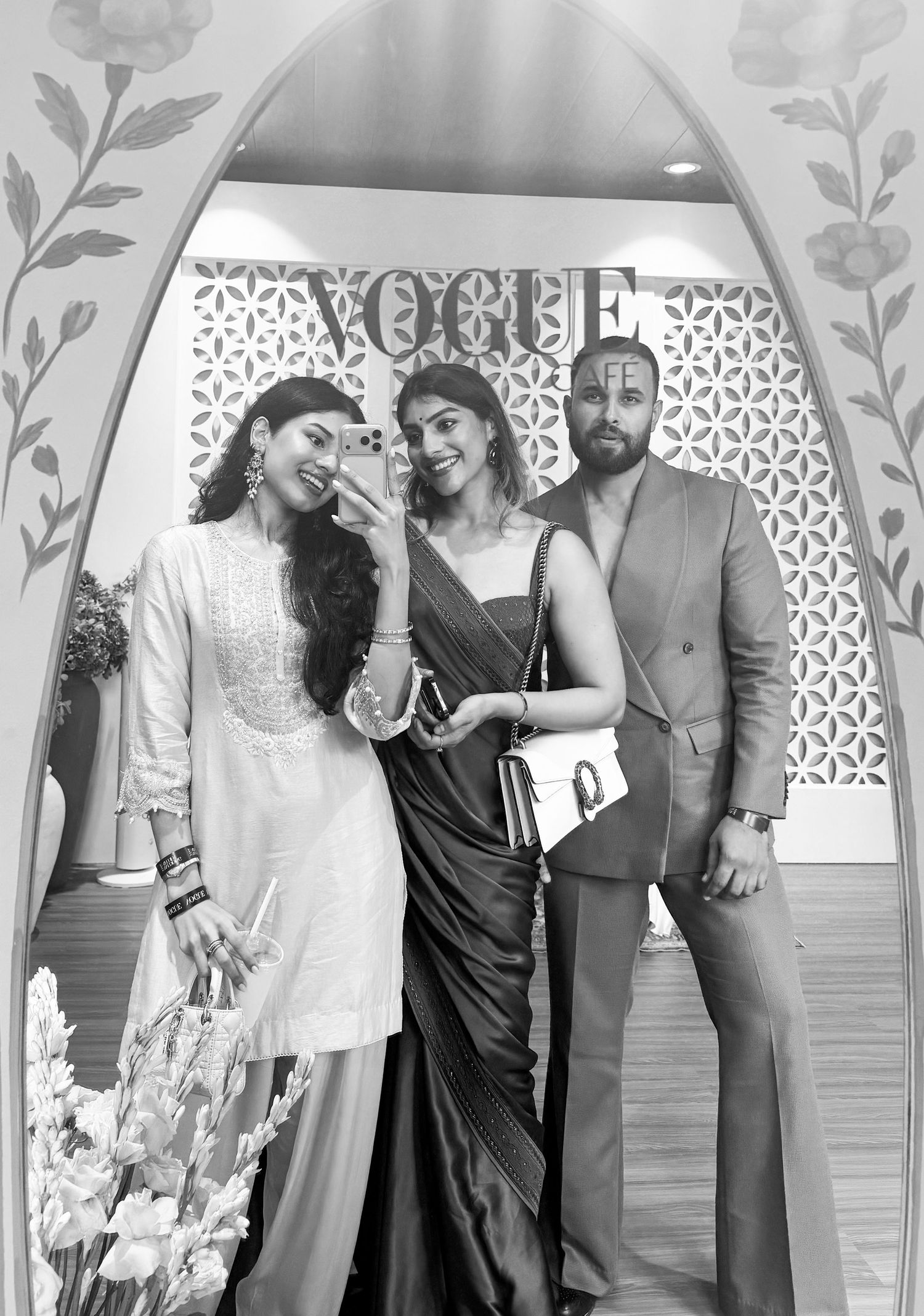 Three elegantly dressed individuals pose for a mirror selfie at a Vogue event in black and white photo.