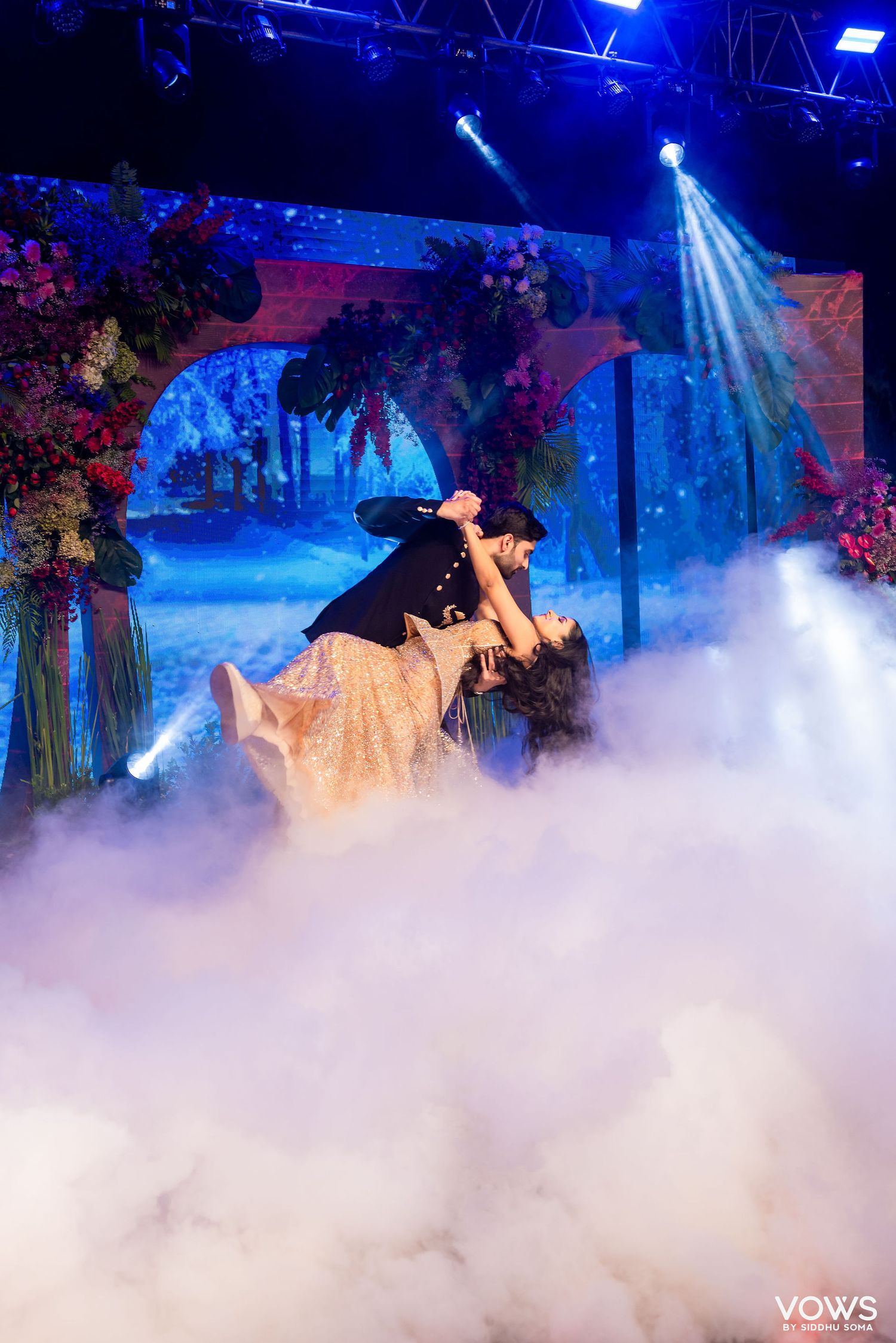 Couple dancing on a fog-filled stage with colorful floral backdrop and dramatic blue stage lighting.