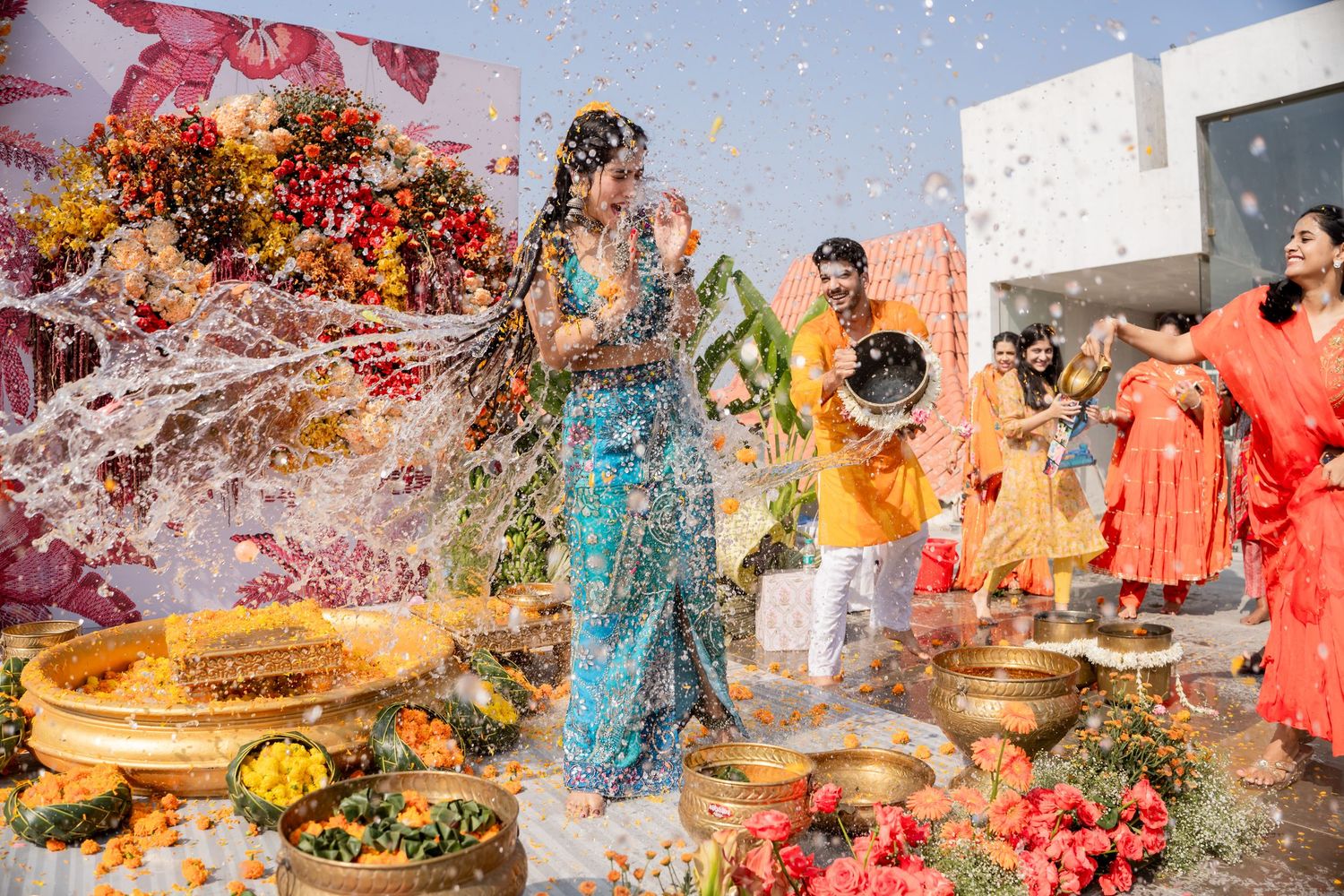 Vibrant Indian wedding Haldi ceremony with guests throwing water and flowers on bride in colorful traditional attire.