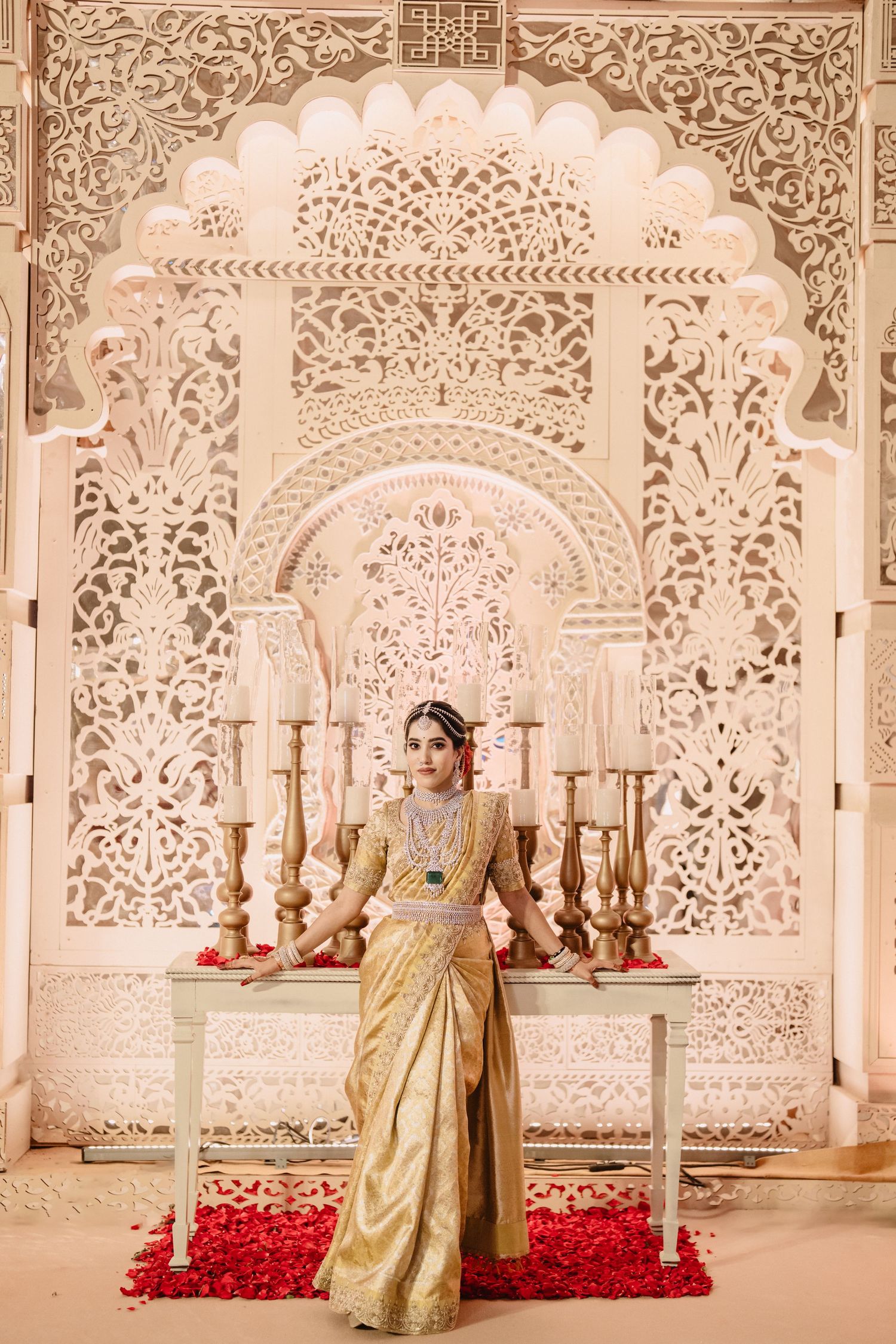 Bride in golden silk saree poses before ornate white carved backdrop with candles and red rose petals.