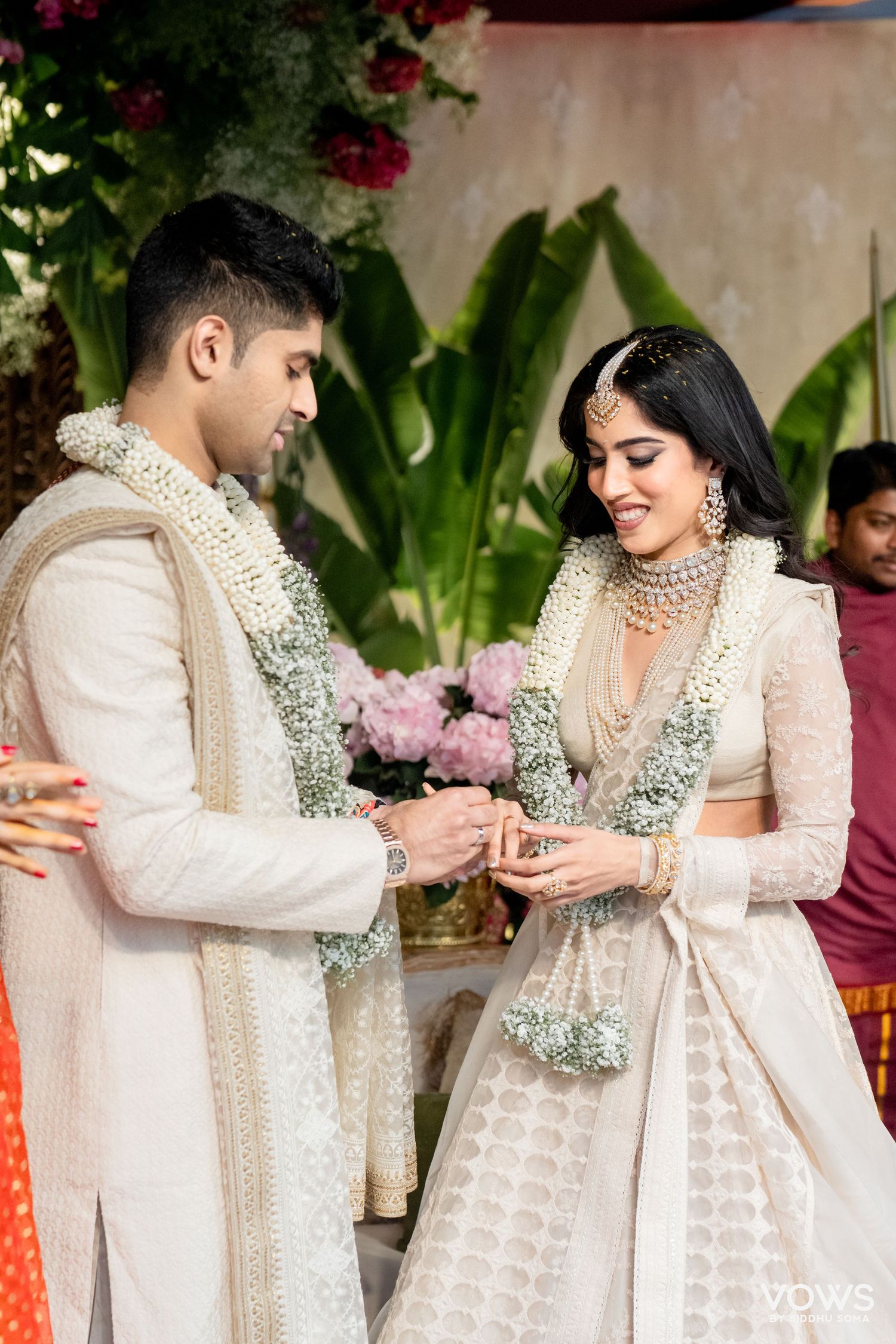 Indian couple in white wedding attire exchange rings during traditional ceremony, adorned with floral garlands.