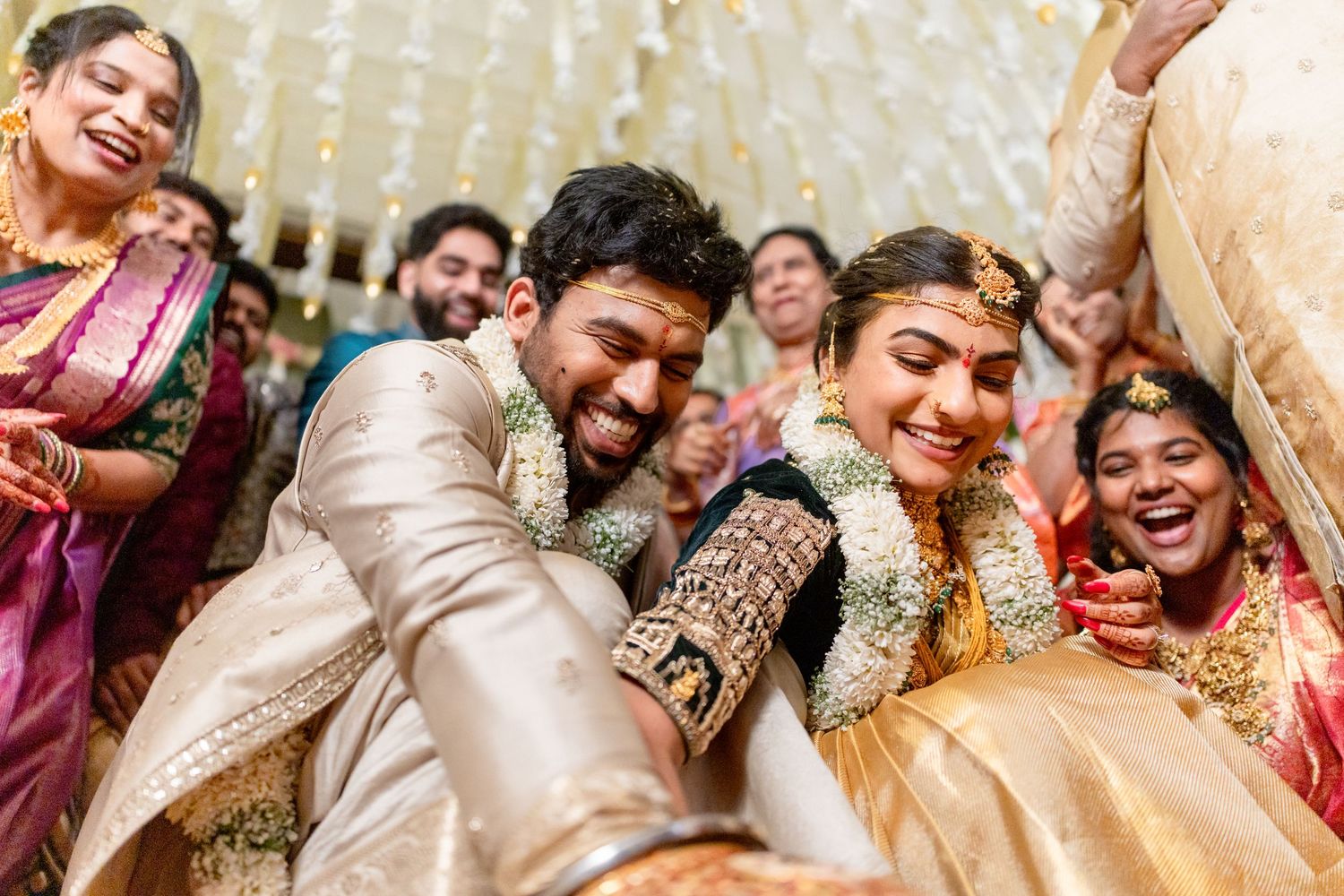 Indian couple in traditional wedding attire sharing a joyful moment surrounded by celebrating guests at a vibrant ceremony.