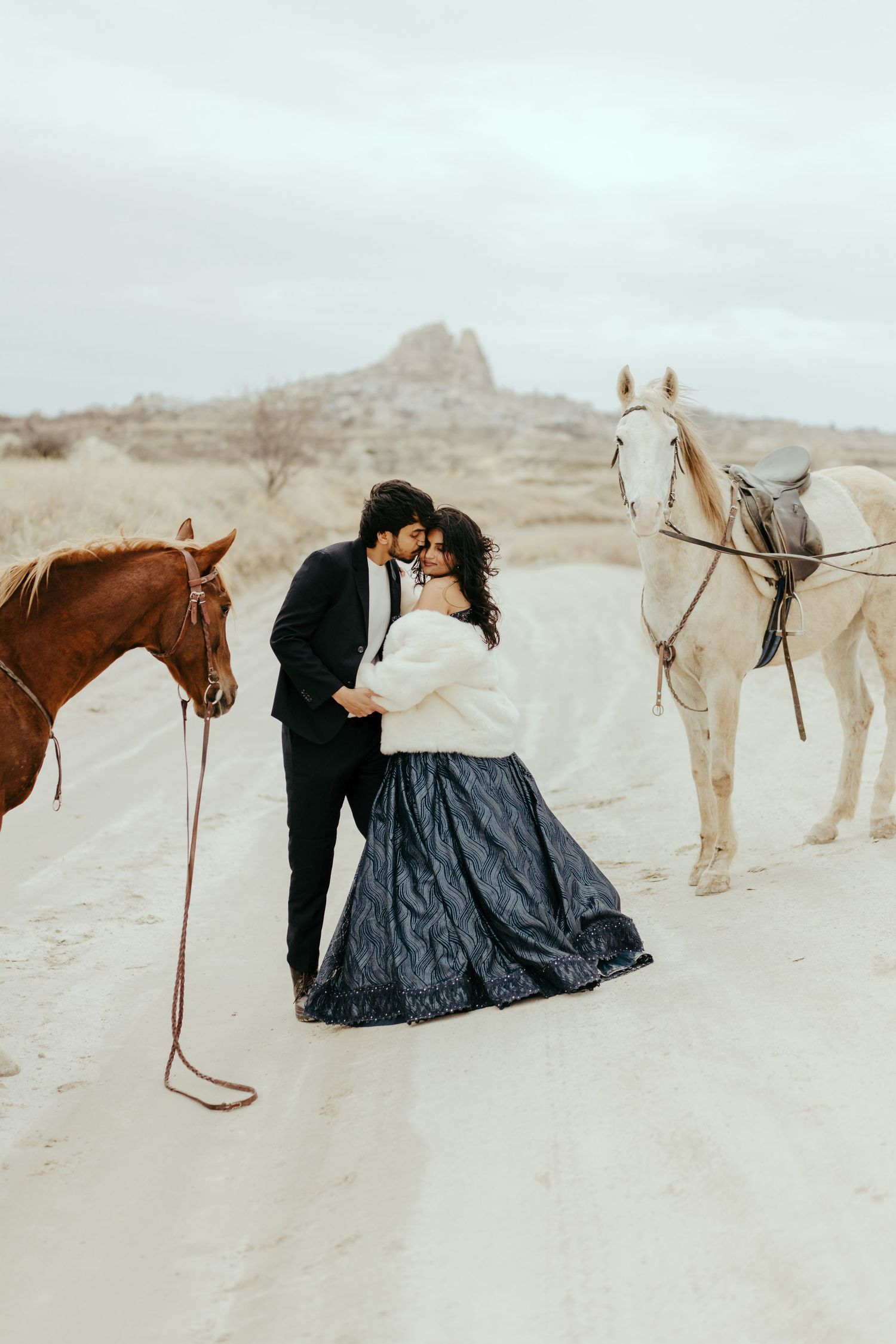 Romantic couple kissing between two horses on a sandy desert path with mountains in the background.