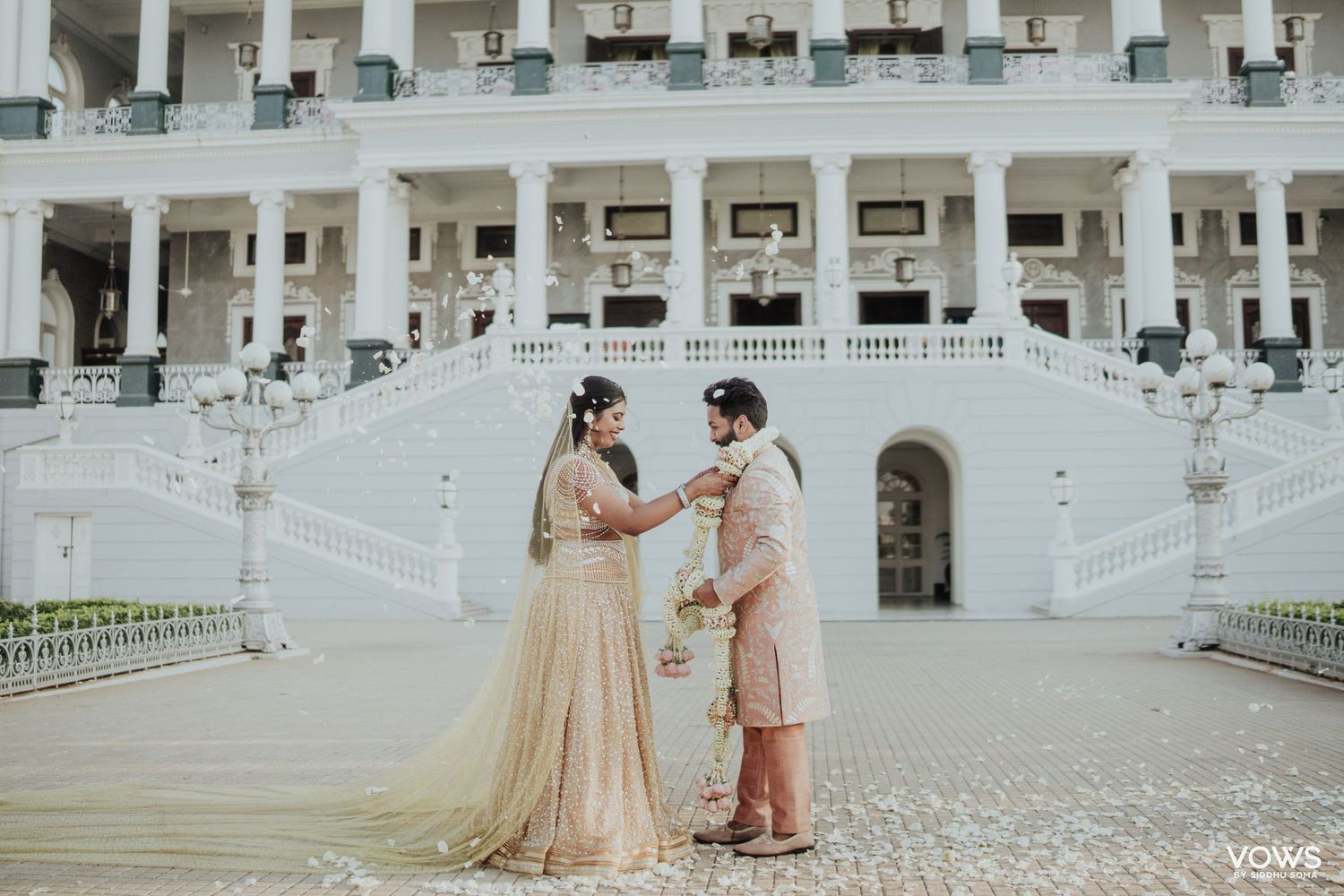 Indian couple in traditional wedding attire exchange garlands at a grand white palace venue.