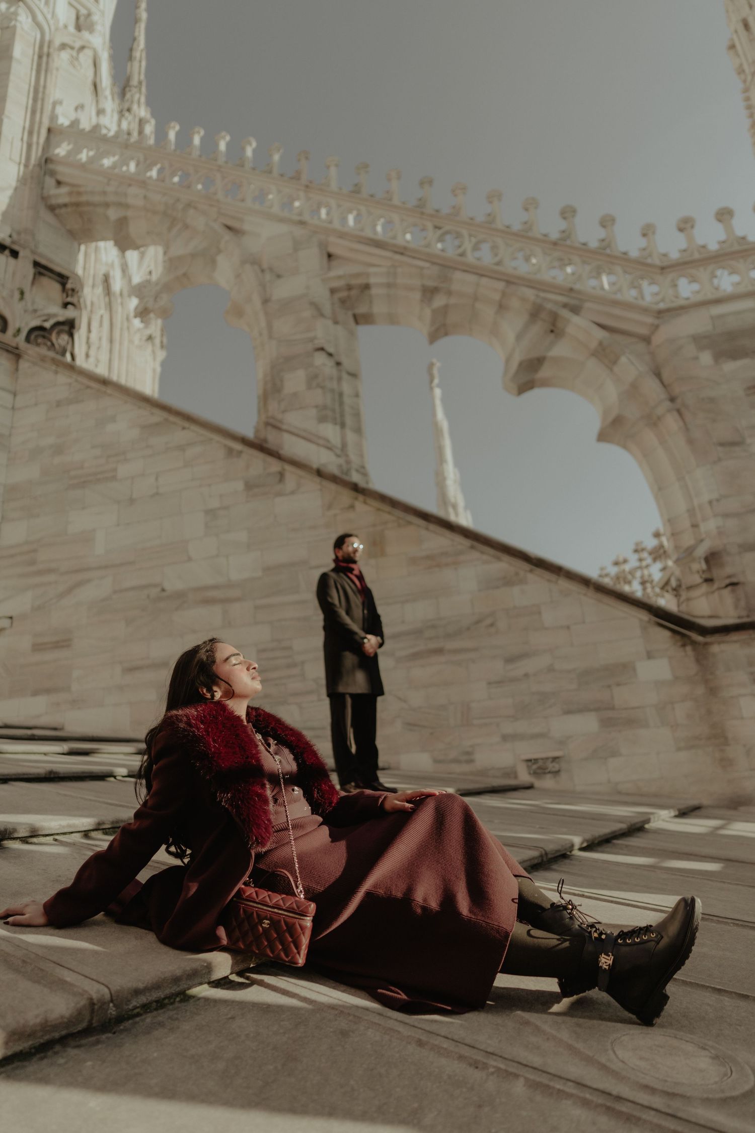 Couple posing dramatically on stone steps of a grand Gothic cathedral, woman in burgundy coat reclines while man stands.