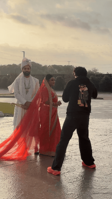 A photographer in dark clothing and red shoes captures a South Asian bride in red and a groom in white at a wet outdoor venue at sunset.
