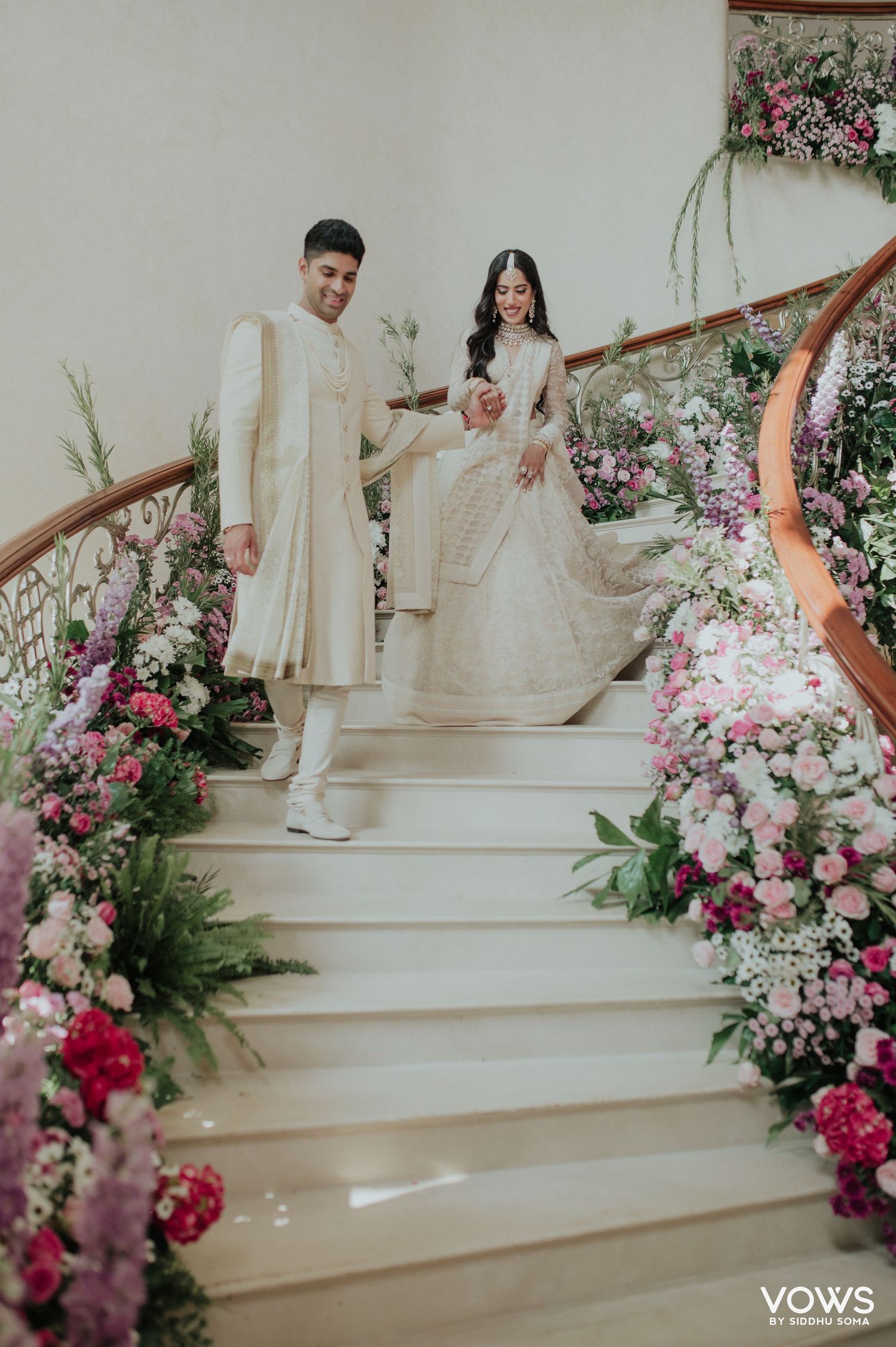 Indian wedding couple in ivory outfits descend flower-lined staircase at elegant venue.