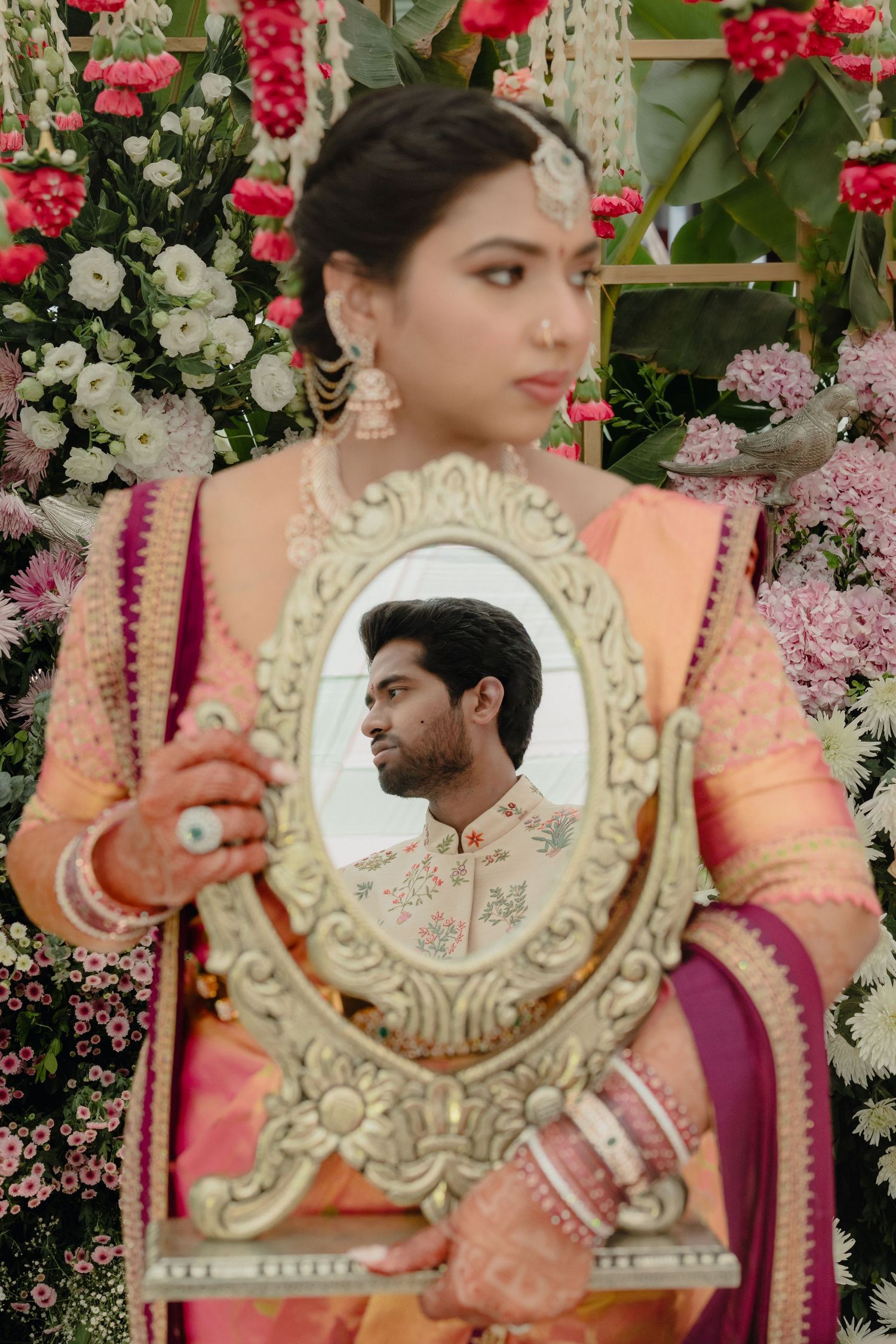 Indian bride in pink saree holds ornate mirror reflecting groom in sherwani, surrounded by floral backdrop.