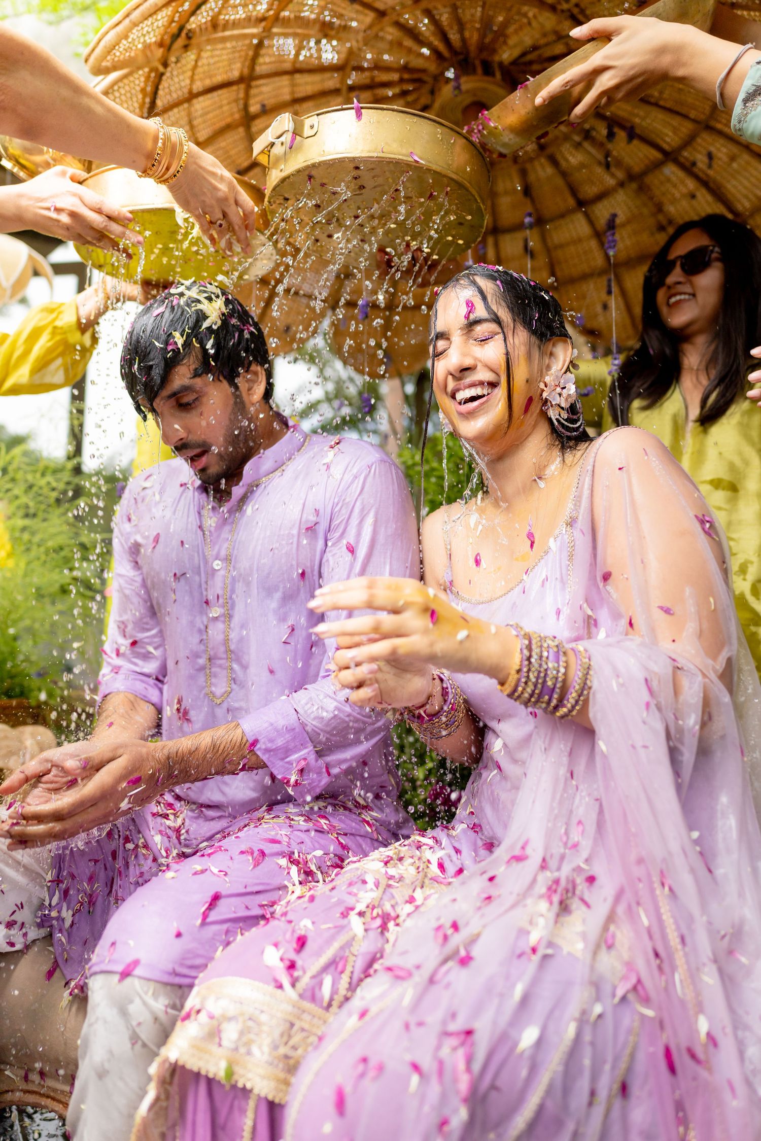 Indian couple in lavender outfits laughing during a floral shower ceremony at a traditional wedding celebration.