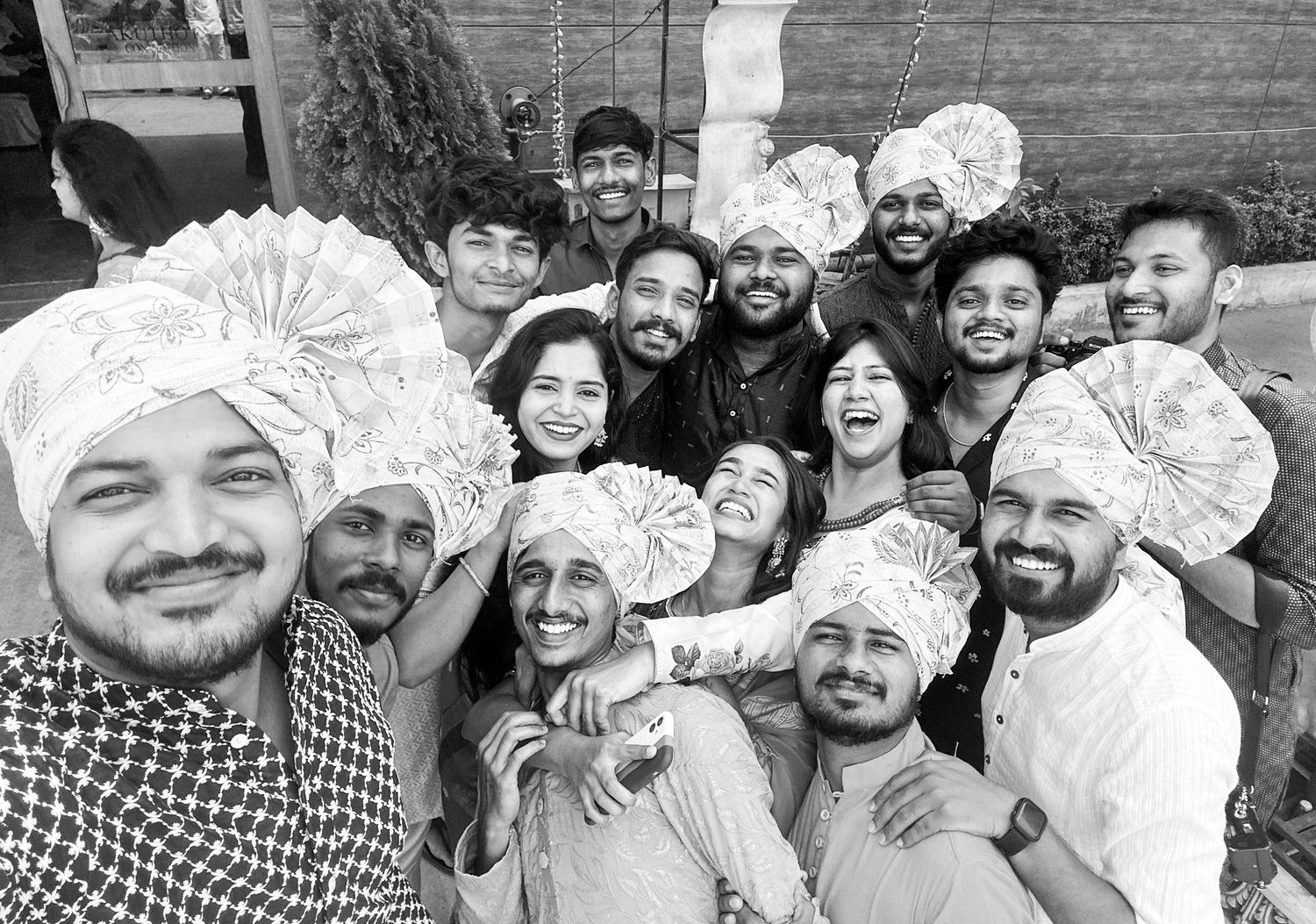 Black and white group selfie of friends celebrating, some wearing decorative turbans, smiling joyfully outdoors.