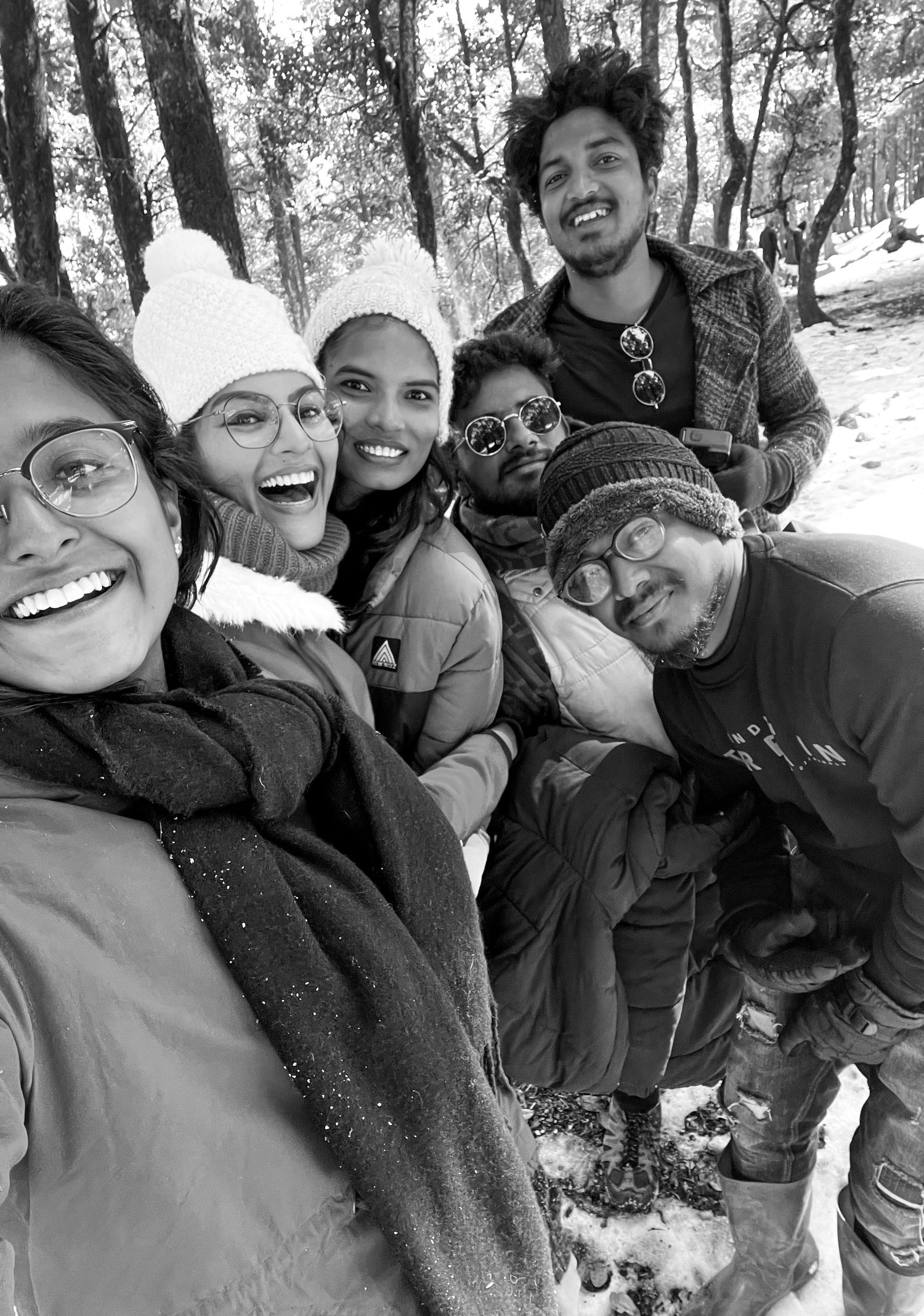 Group of friends smiling together in a snowy forest, wearing winter hats and jackets in a black and white selfie.