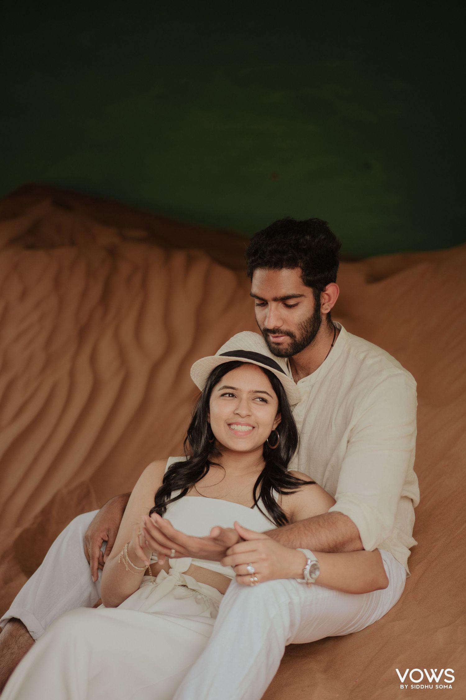 Couple sitting on red sand dunes, woman in white dress and hat, man in cream outfit, romantic desert photoshoot.