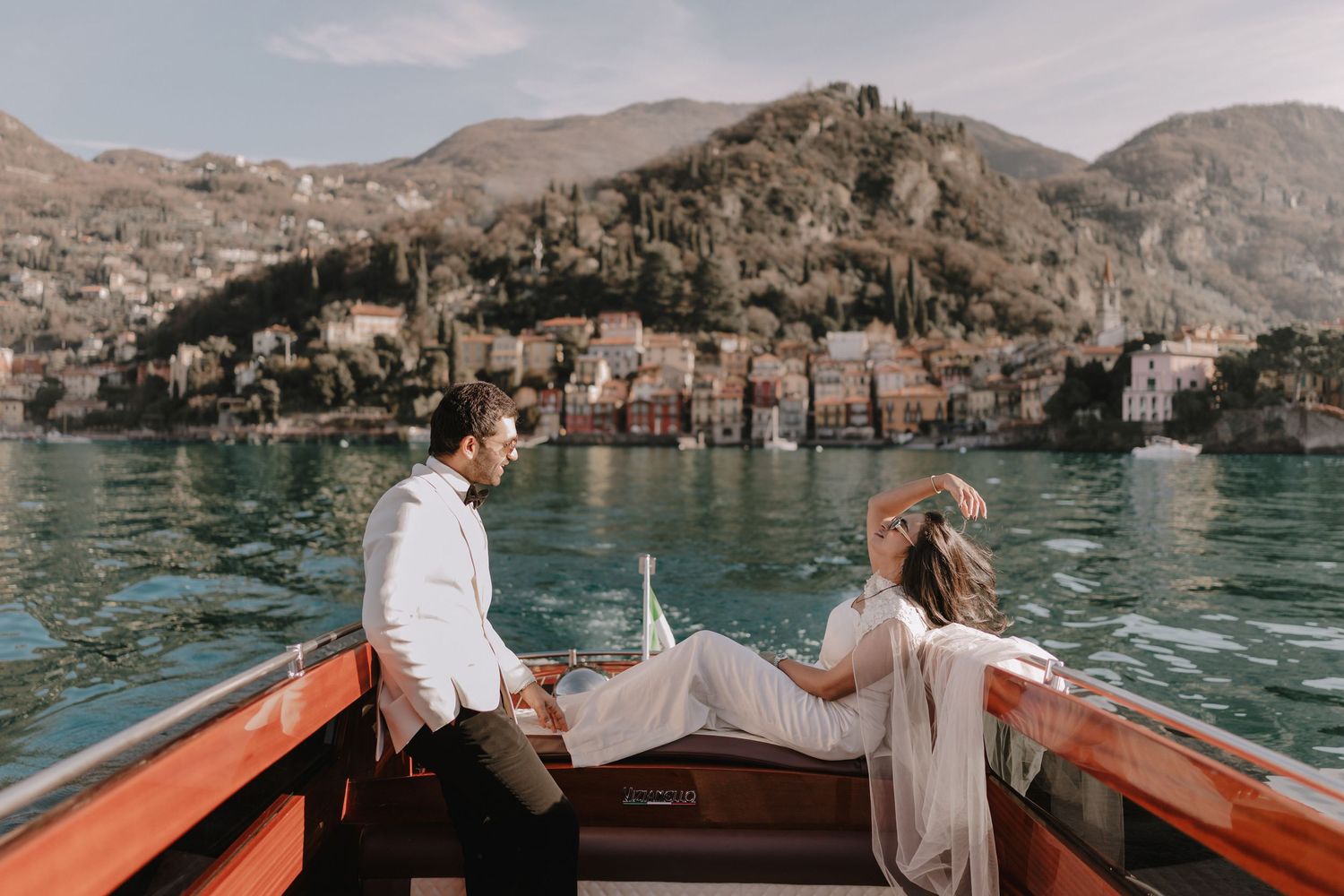 Bride and groom in wedding attire relax on a classic wooden boat with a charming lakeside village and mountains in the background.