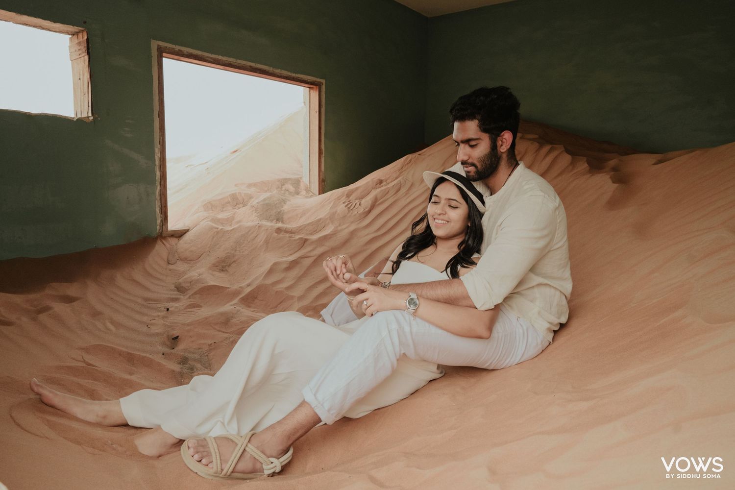 Couple in white outfits sitting romantically on sand dunes inside a room with olive green walls.