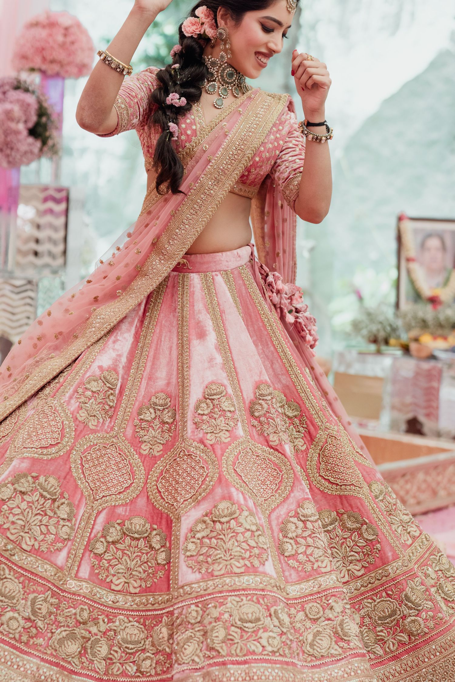 Bride in pink embroidered lehenga with gold zari work and dupatta, wearing floral hair accessories at wedding.