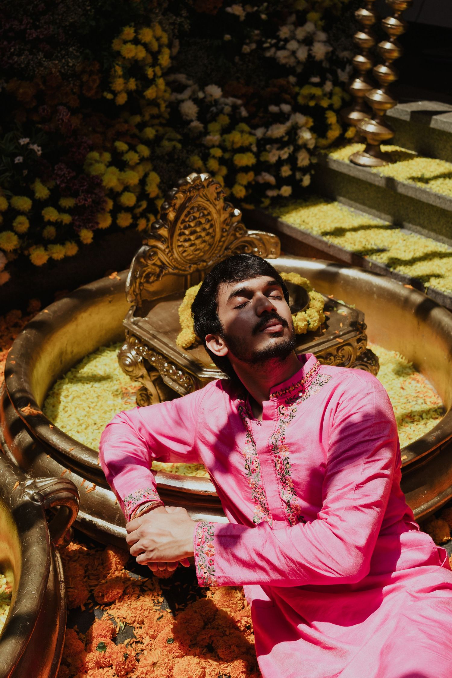 Man in pink embroidered kurta relaxing in ornate golden setting surrounded by yellow flowers.