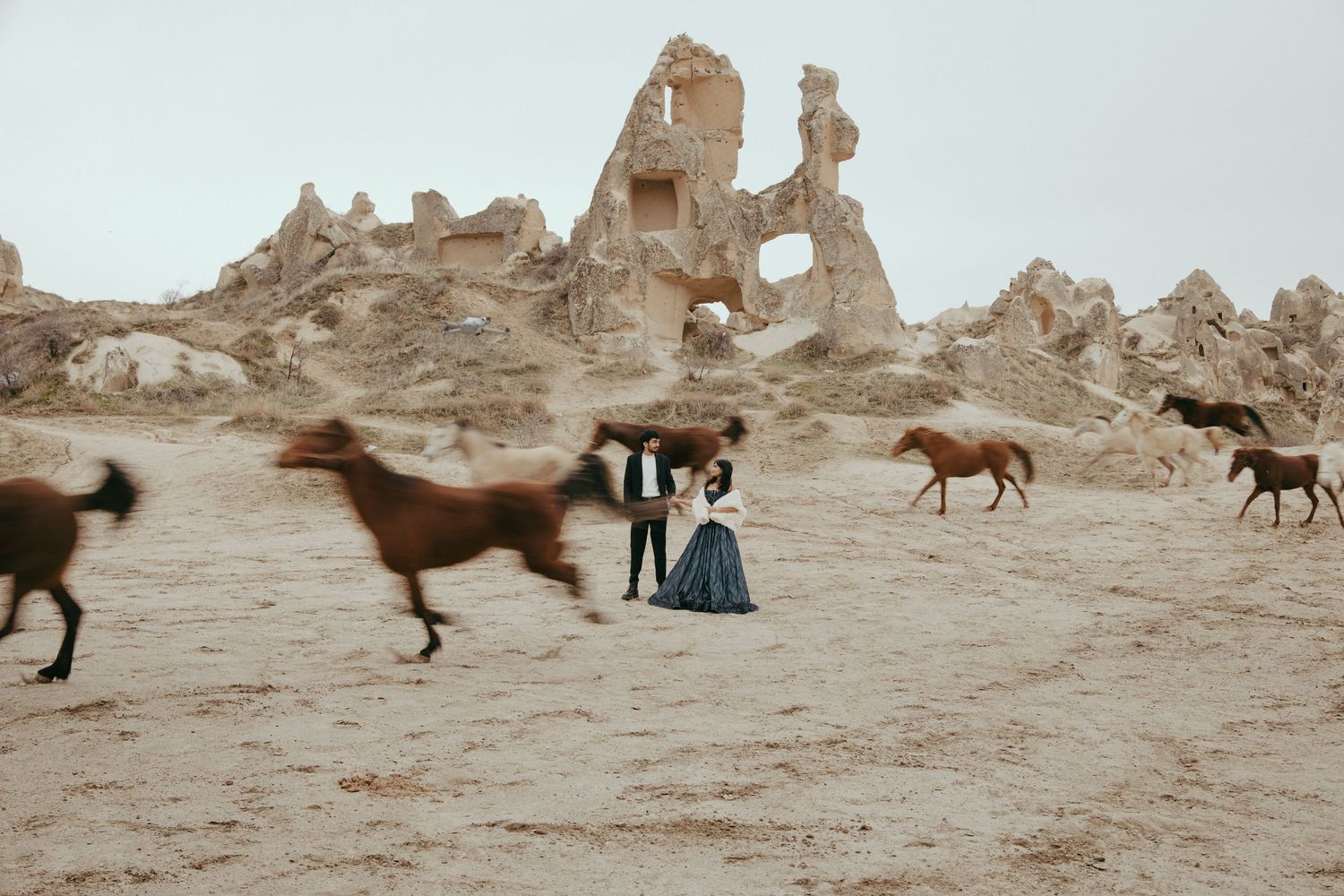 Couple in formal attire poses among wild horses and ancient rock formations in a desert landscape.